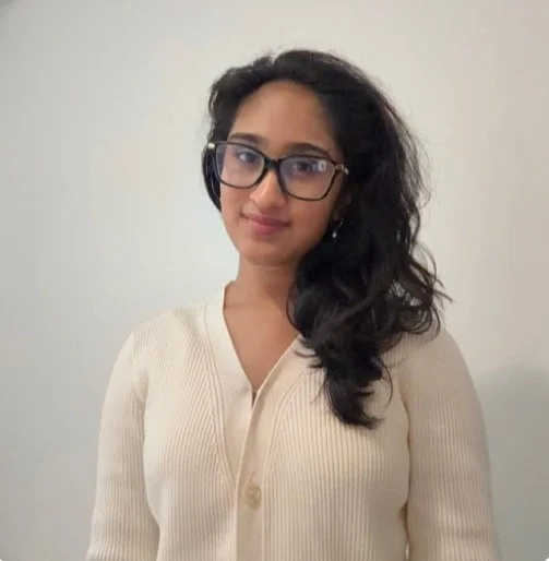 A young woman with long, wavy black hair, wearing glasses and a cream-colored cardigan, standing in front of a plain off-white wall.