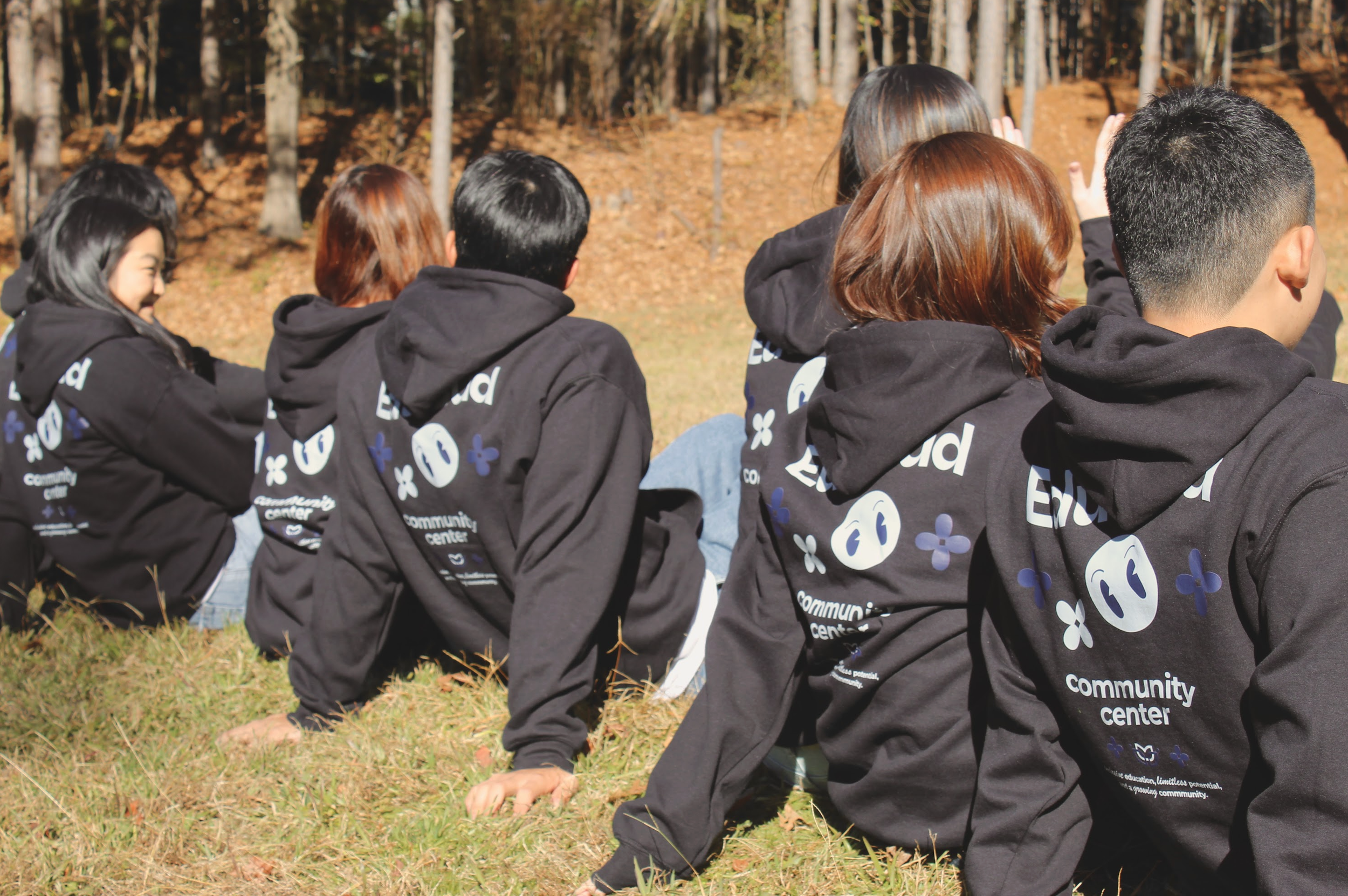 A group of people sitting outdoors in autumn, wearing matching black hoodies with text and graphics, with trees and fallen leaves in the background.
