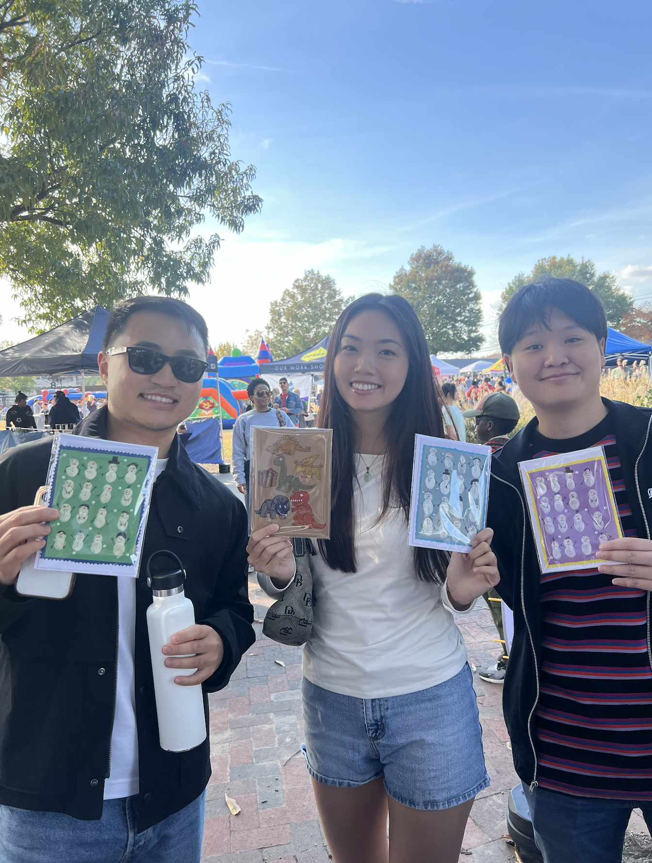 Three smiling people holding colorful holiday-themed stickers at an outdoor event with booths and inflatable rides in the background.
