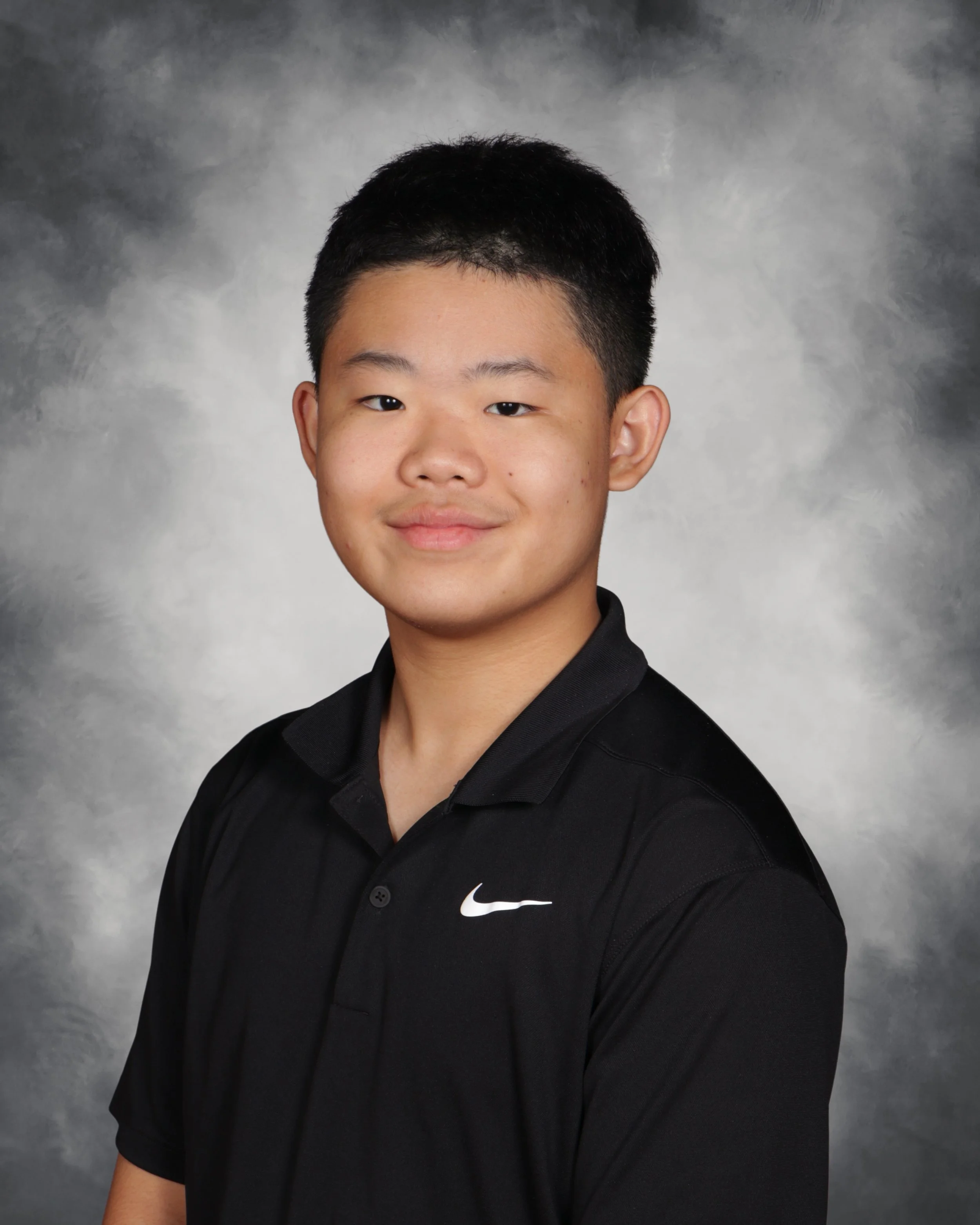 A young male student with short black hair and a slight smile, wearing a black Nike polo shirt, standing against a gray, textured background.