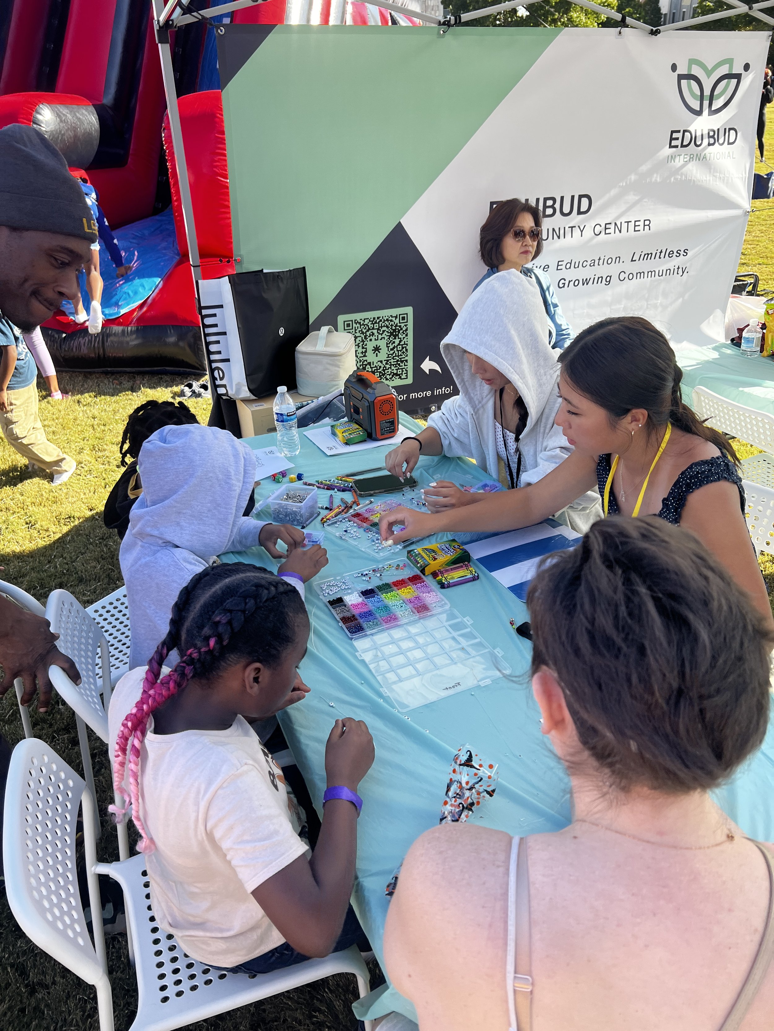 A group of children and adults gathered around a table at an outdoor community event, engaging in arts and crafts, with a large banner in the background that reads 'EDUBUB Community Center'.
