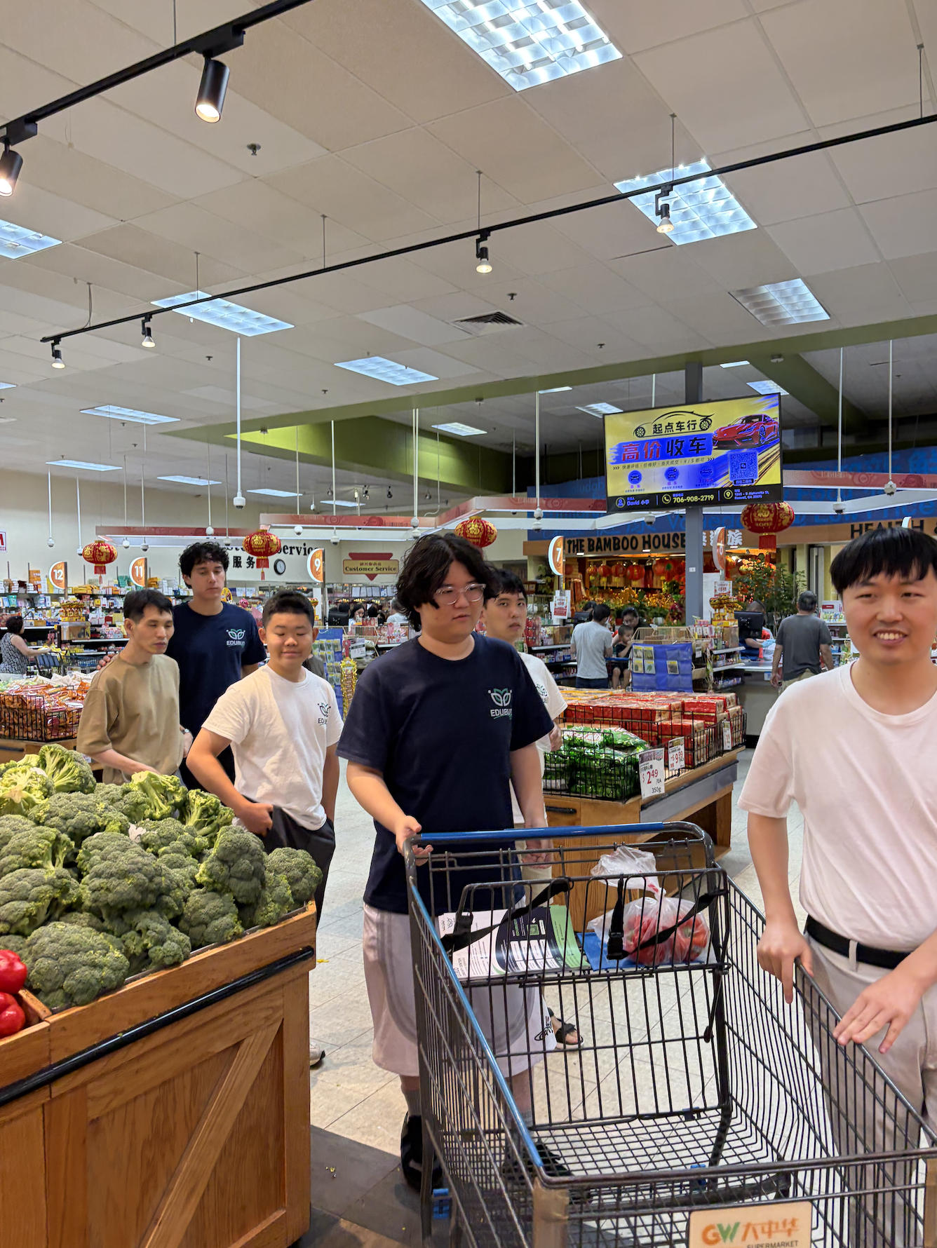 A group of young people with shopping carts in a grocery store aisle, with broccoli and other produce visible in the foreground and signs in Chinese characters overhead.