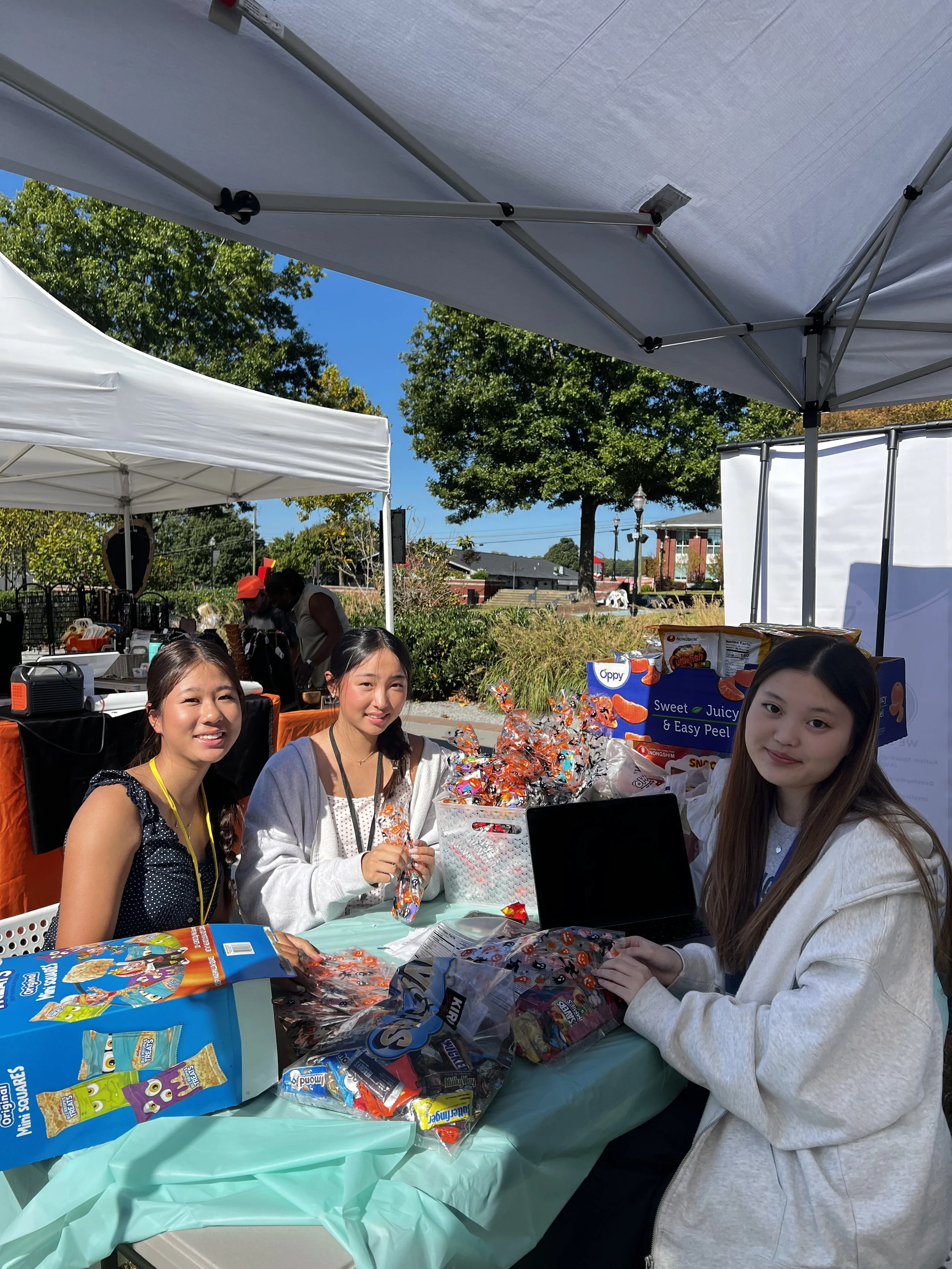 Three women sitting at a table under a white canopy tent at an outdoor event, surrounded by various snacks and candies, with trees and blue sky in the background.