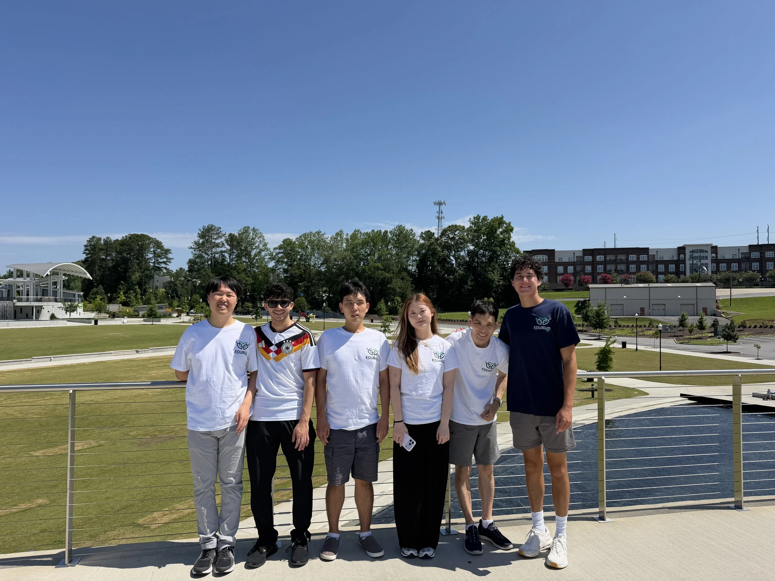 A group of seven young people standing outdoors on a sunny day, smiling and posing for a photo, with green trees, a blue sky, and buildings in the background.