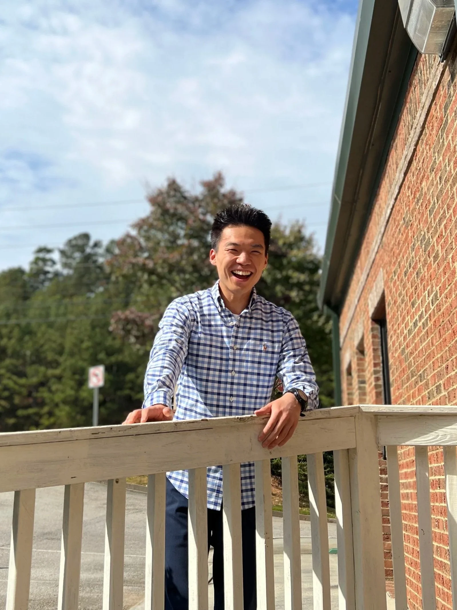 A young man in a blue and white checkered shirt smiling and leaning on a wooden fence, with a background of trees and a brick building, under a partly cloudy sky.