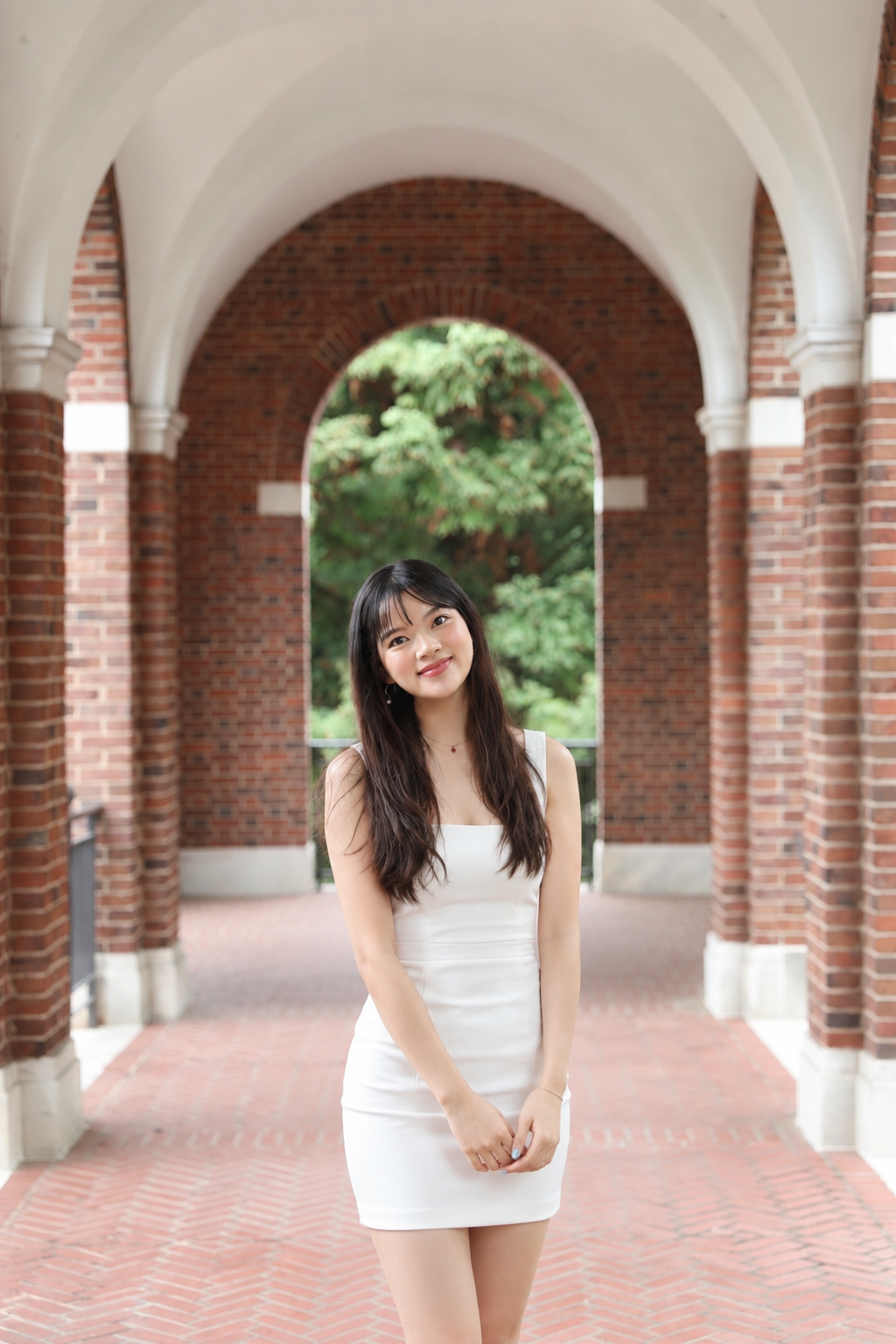 Young woman in white dress standing in an arched brick walkway with greenery in the background.