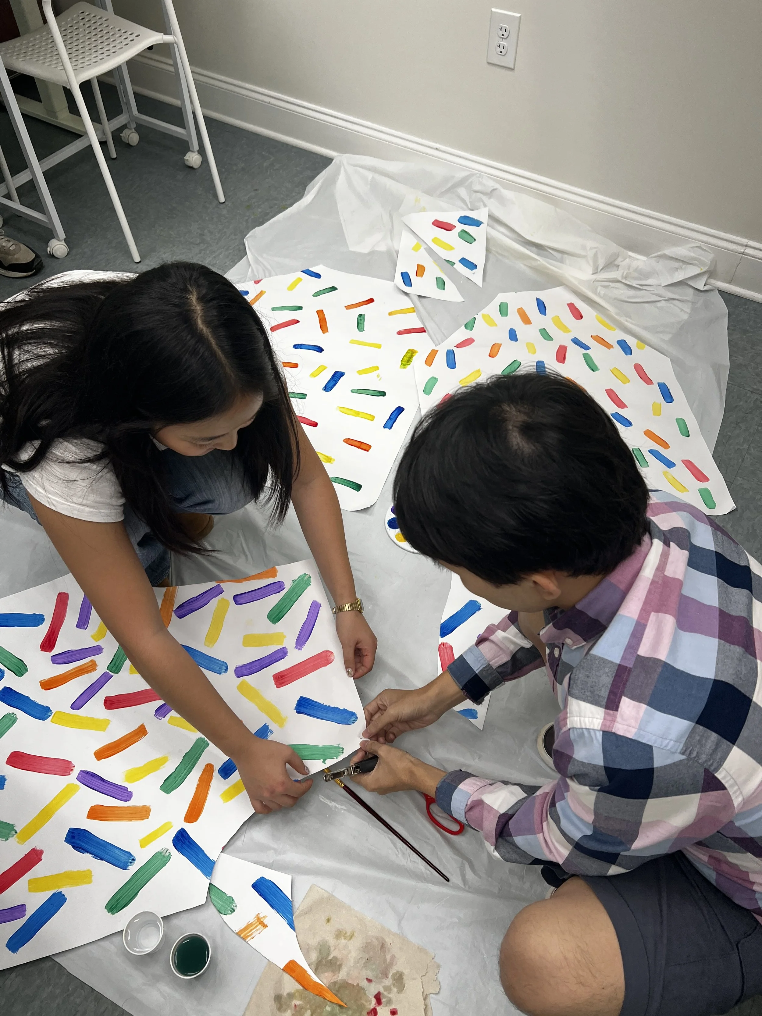 Two people are creating artwork on large sheets of white paper, using colorful paint strokes in red, yellow, green, blue, purple, and orange. They are seated on the floor, with painting supplies like paint cups, brushes, and paper towels nearby. One 