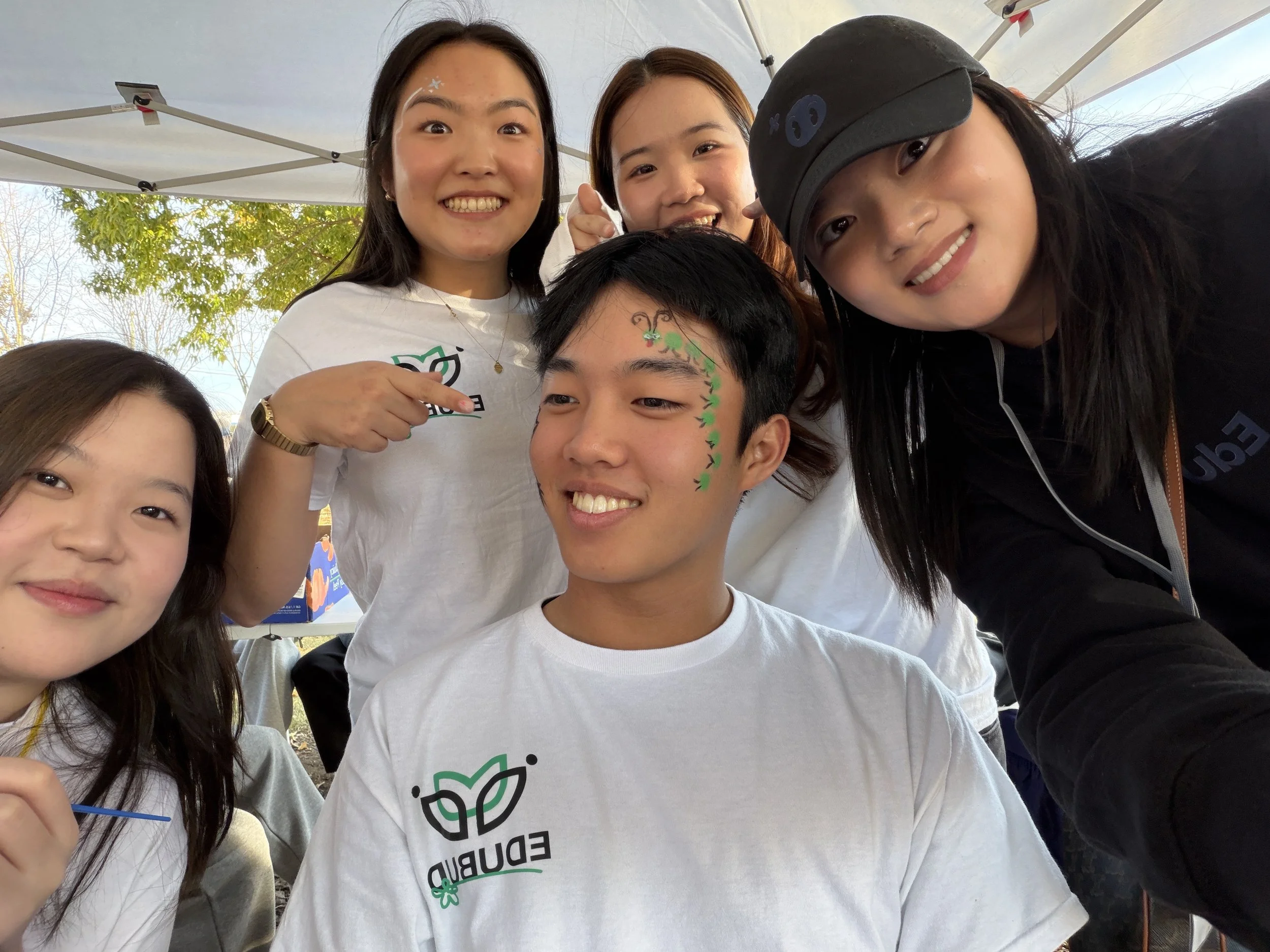 A group of young people smiling and taking a selfie outdoors under a canopy, with trees in the background, during what appears to be a community or environmental event.