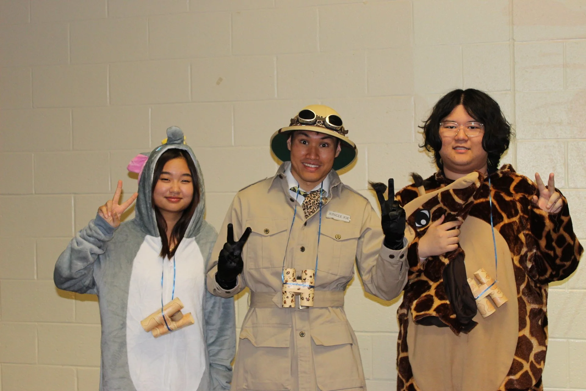 Three people dressed in animal and explorer costumes posing with peace signs against a beige wall.