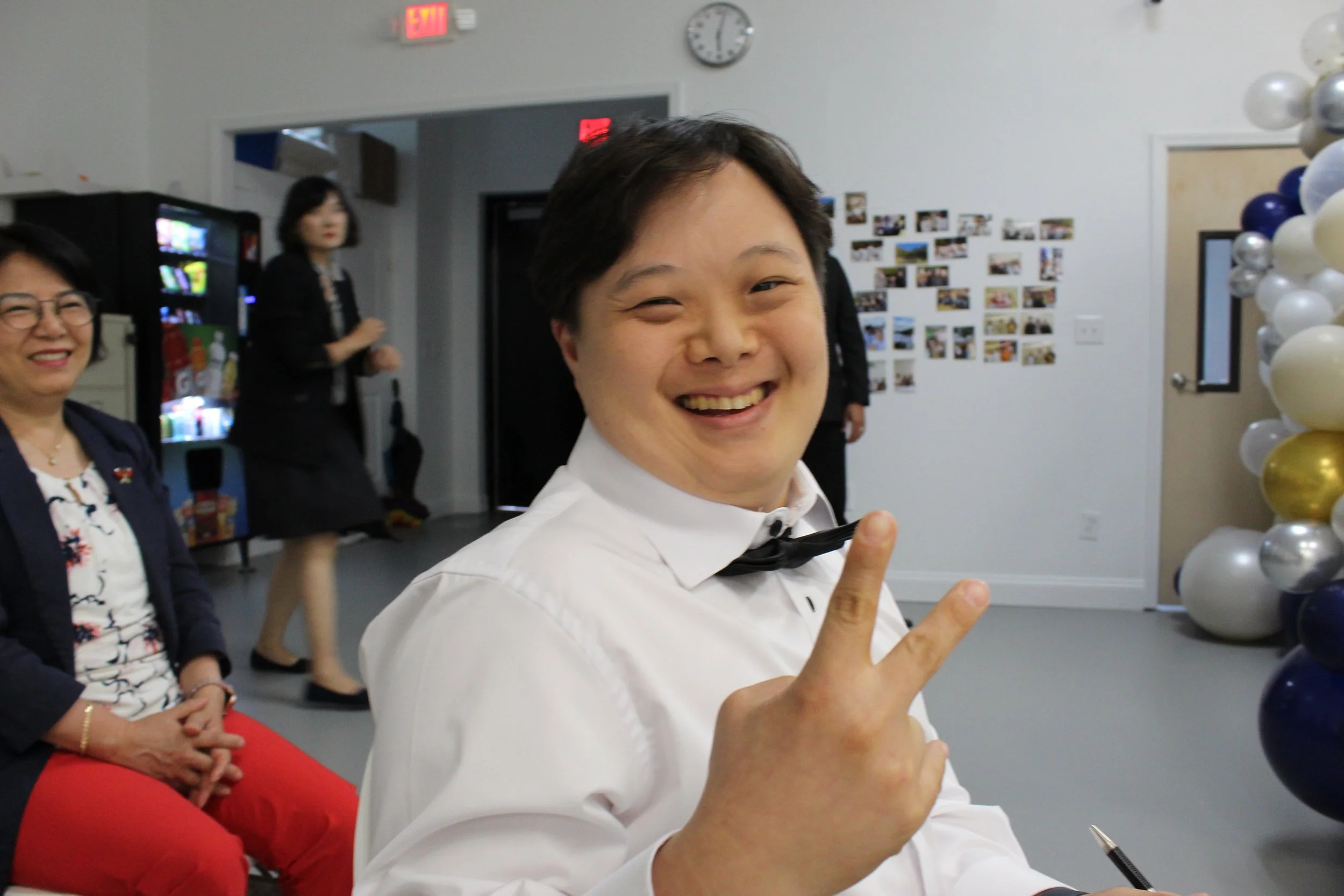 A smiling person in a white shirt and black bowtie making a peace sign with their fingers at an indoor event. In the background, there are two women, one smiling and the other slightly blurred, seated near a vending machine and decorated walls with p