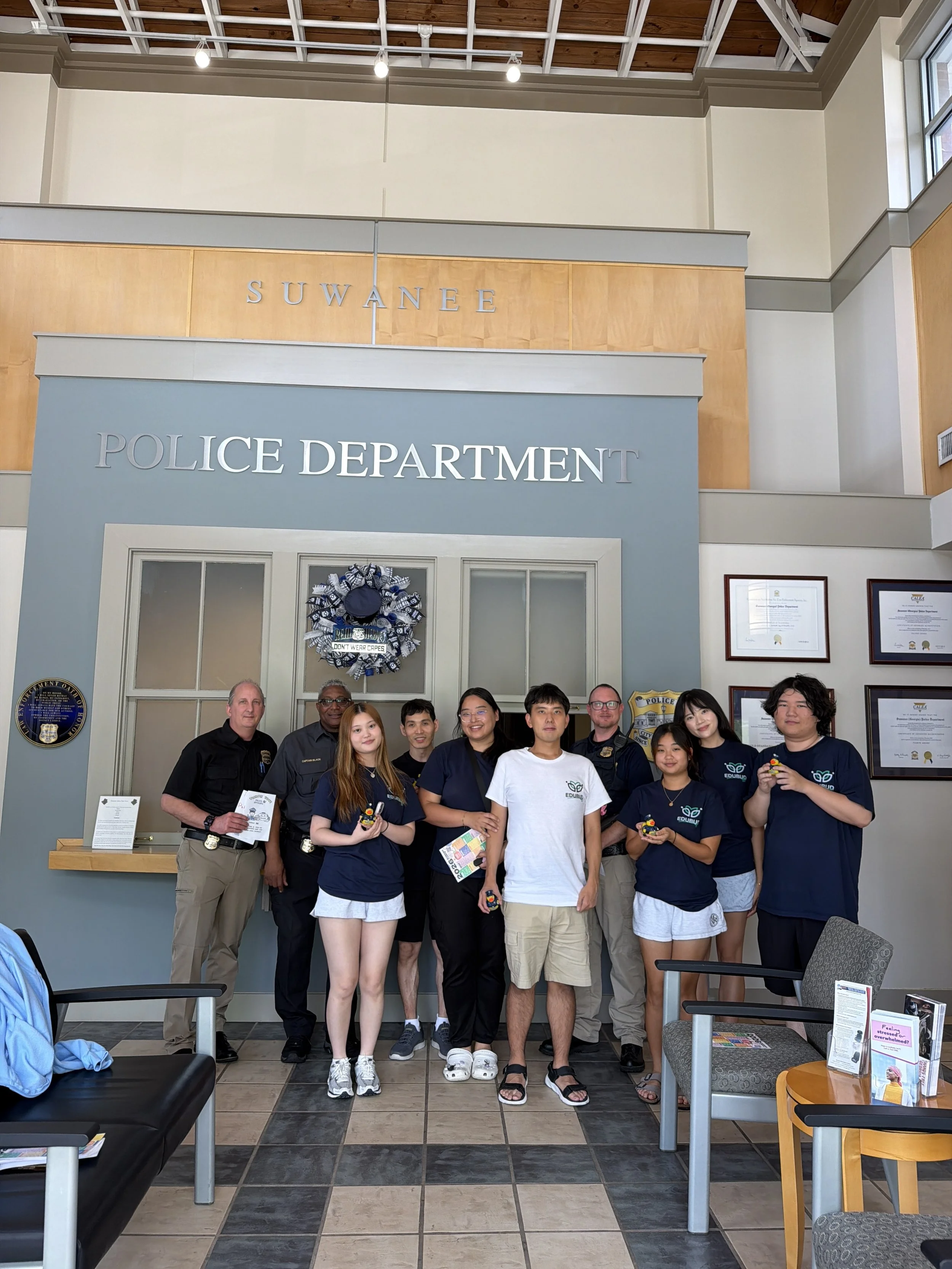 Group of people standing inside a police department, with a sign that says 'POLICE DEPARTMENT' above them. The group includes children and adults, some in uniforms and others in casual clothing, posing for a photo.