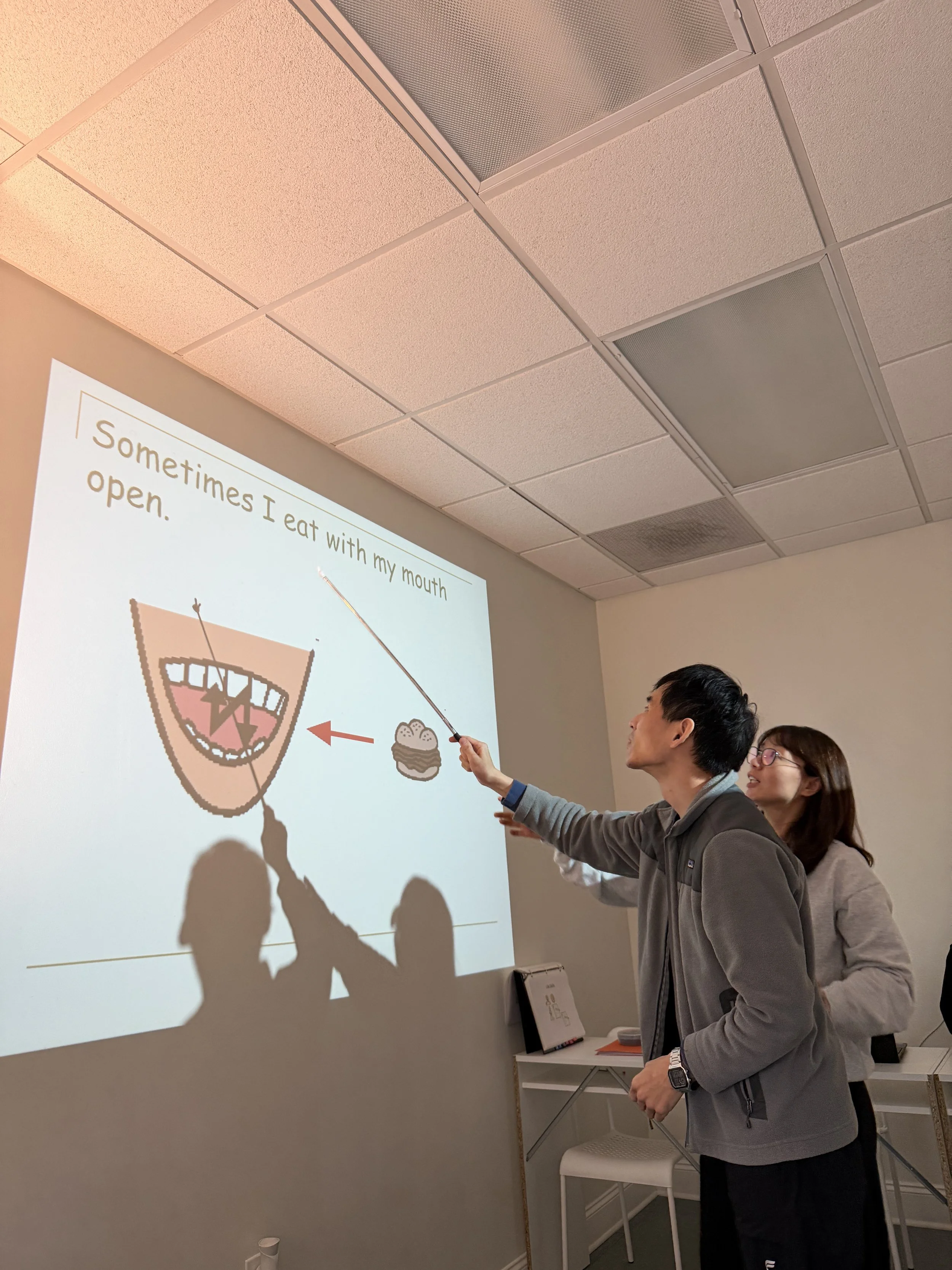 Two people standing in a room, giving a presentation projected on the wall, which features a cartoonish diagram with a mouth, teeth, and an arrow pointing to a cupcake, with a caption about eating with their mouth.
