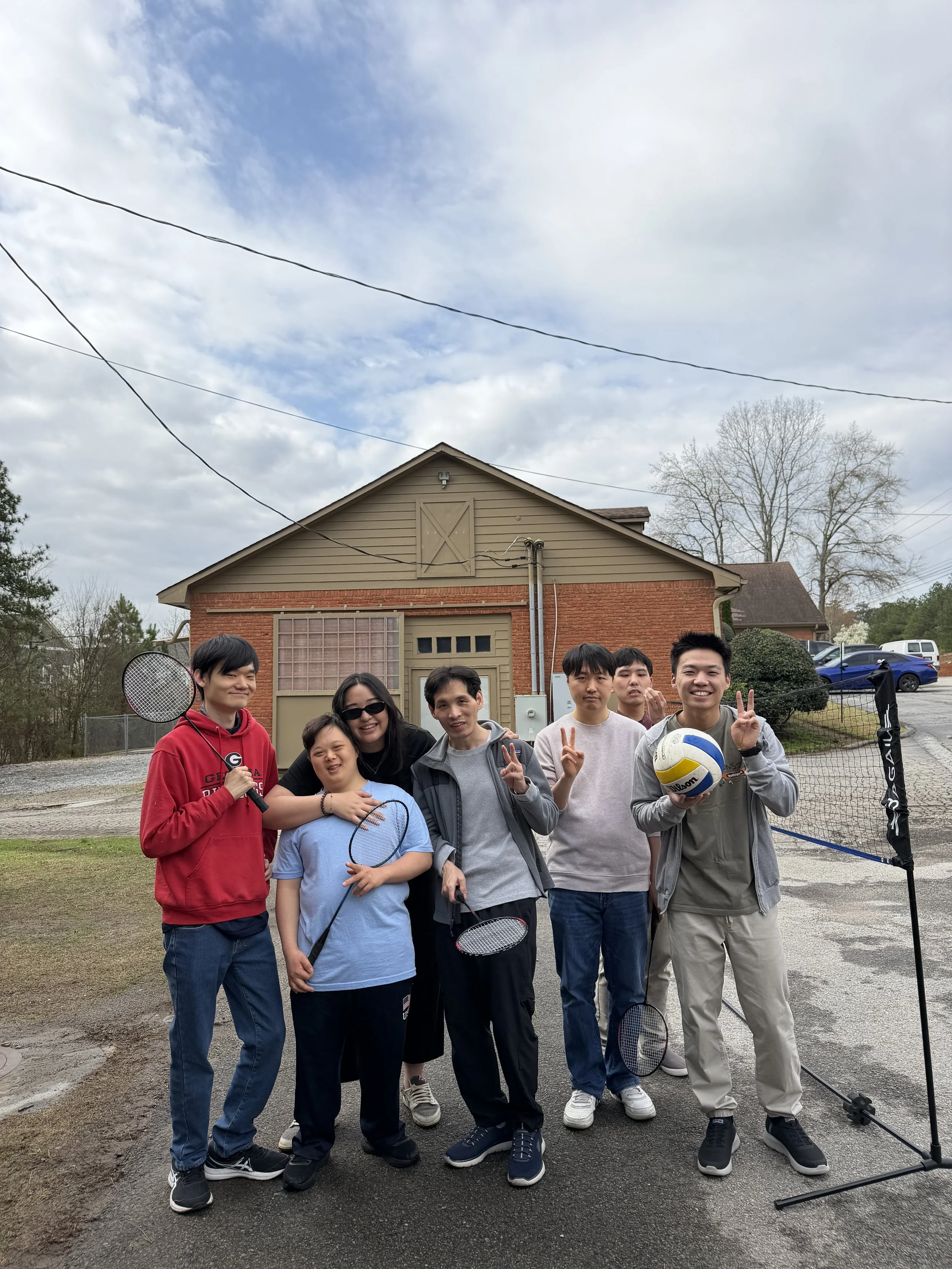 Group of eight young people standing outdoors in front of a house, holding badminton rackets and a volleyball, smiling and making peace signs.