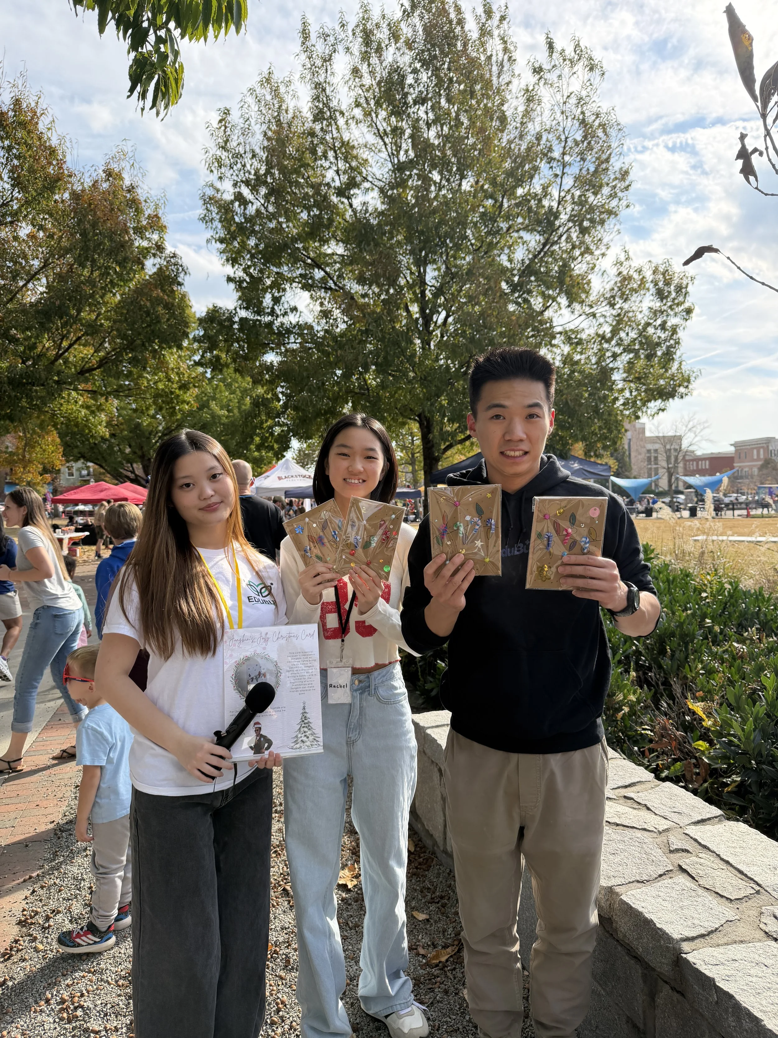 Three young people holding decorated Christmas cards outdoors at a festive event, with other people, tents, and trees in the background.