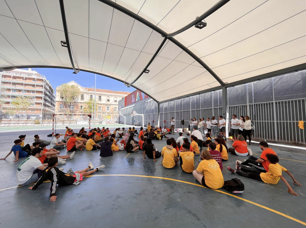 A large group of children and teenagers sitting on the ground inside a semi-covered outdoor sports court, listening to a speaker or instructor at the front. Some are wearing orange shirts, and the court is surrounded by tall buildings.