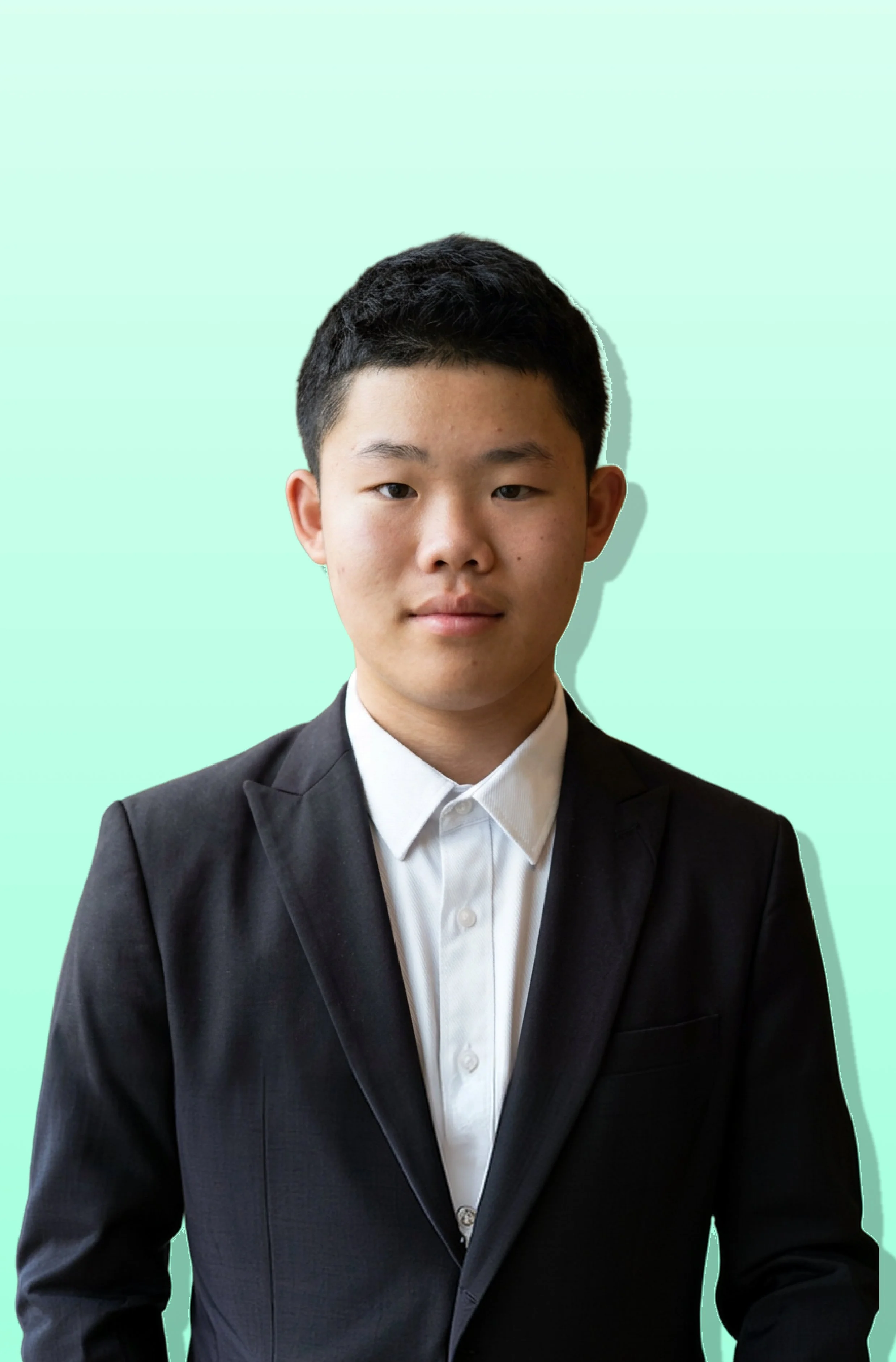 A young man with short dark hair wearing a black suit and white dress shirt, sitting against a light green background.