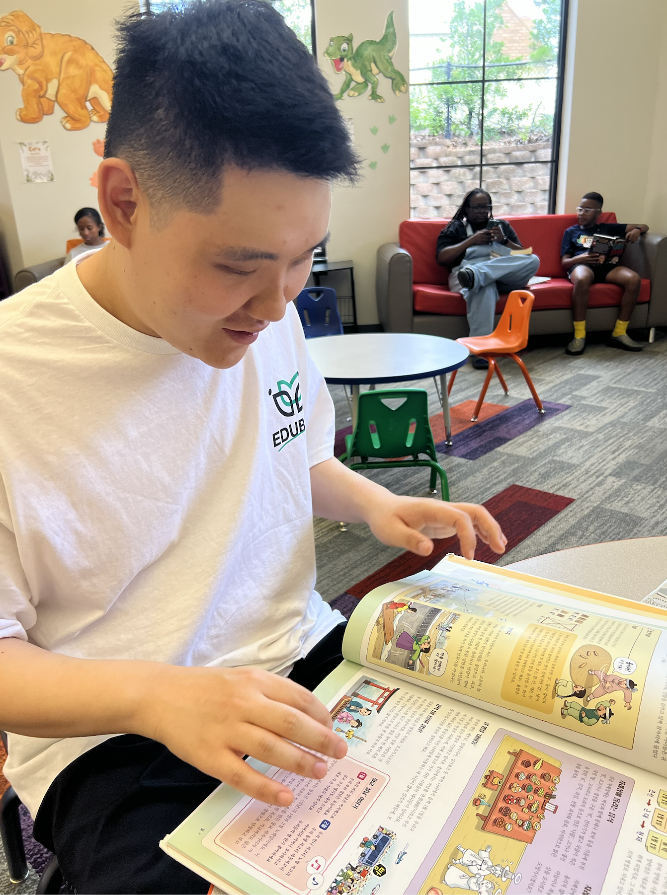 A young man with short black hair is reading a colorful comic book or magazine inside a room. In the background, two children are sitting on a red couch, one with a tablet and the other reading a book. The room has cartoon animal wall decorations and