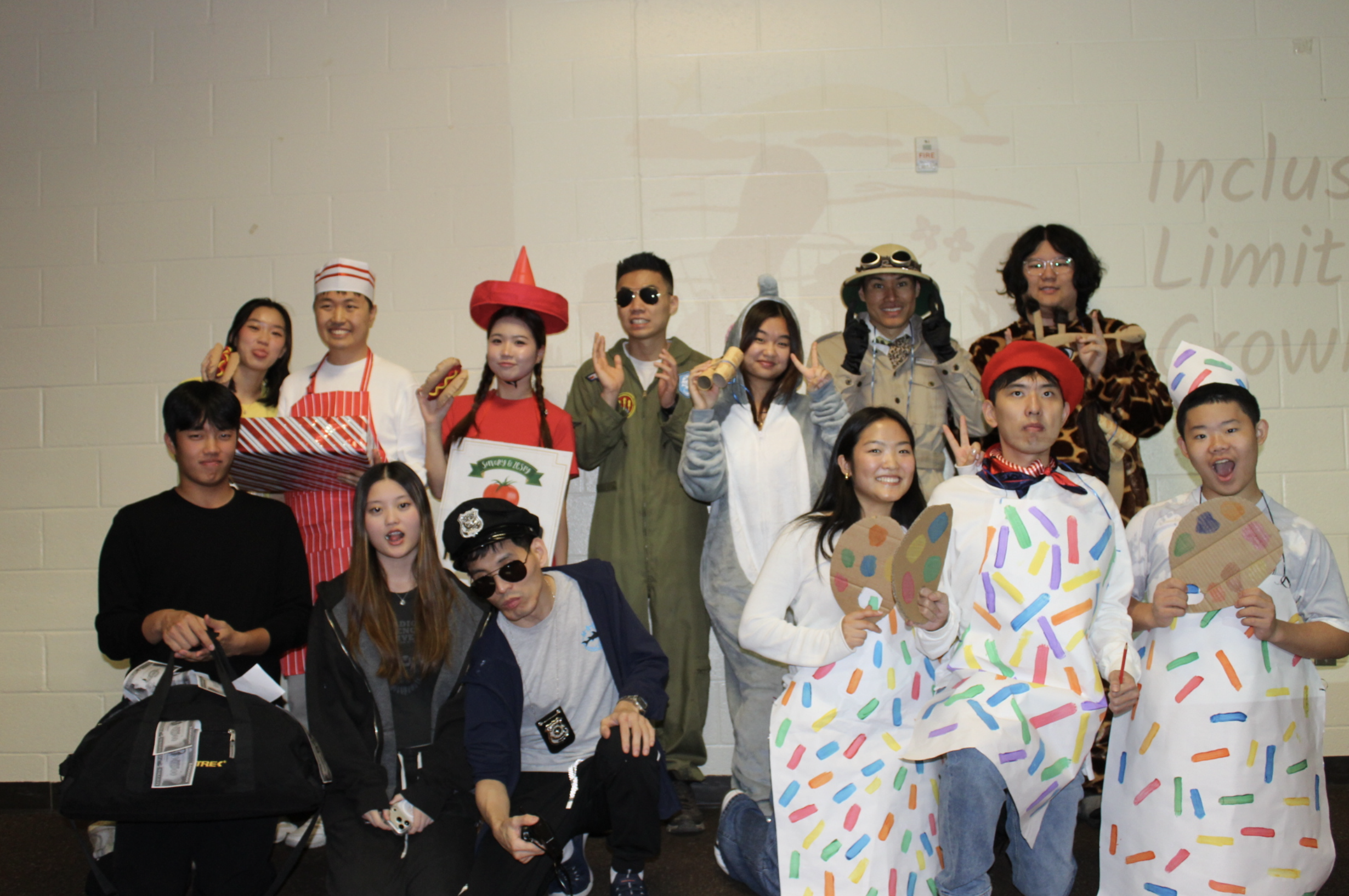 Group of people in costumes, some holding props, standing indoors against a light-colored wall.