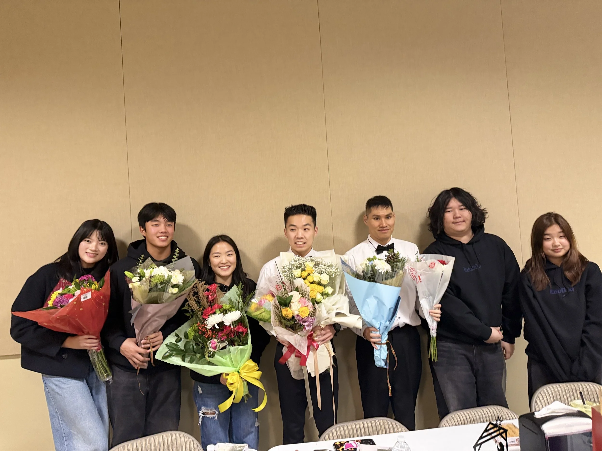Group of young people standing indoors, holding bouquets of flowers, smiling for a photo at a celebration or event.
