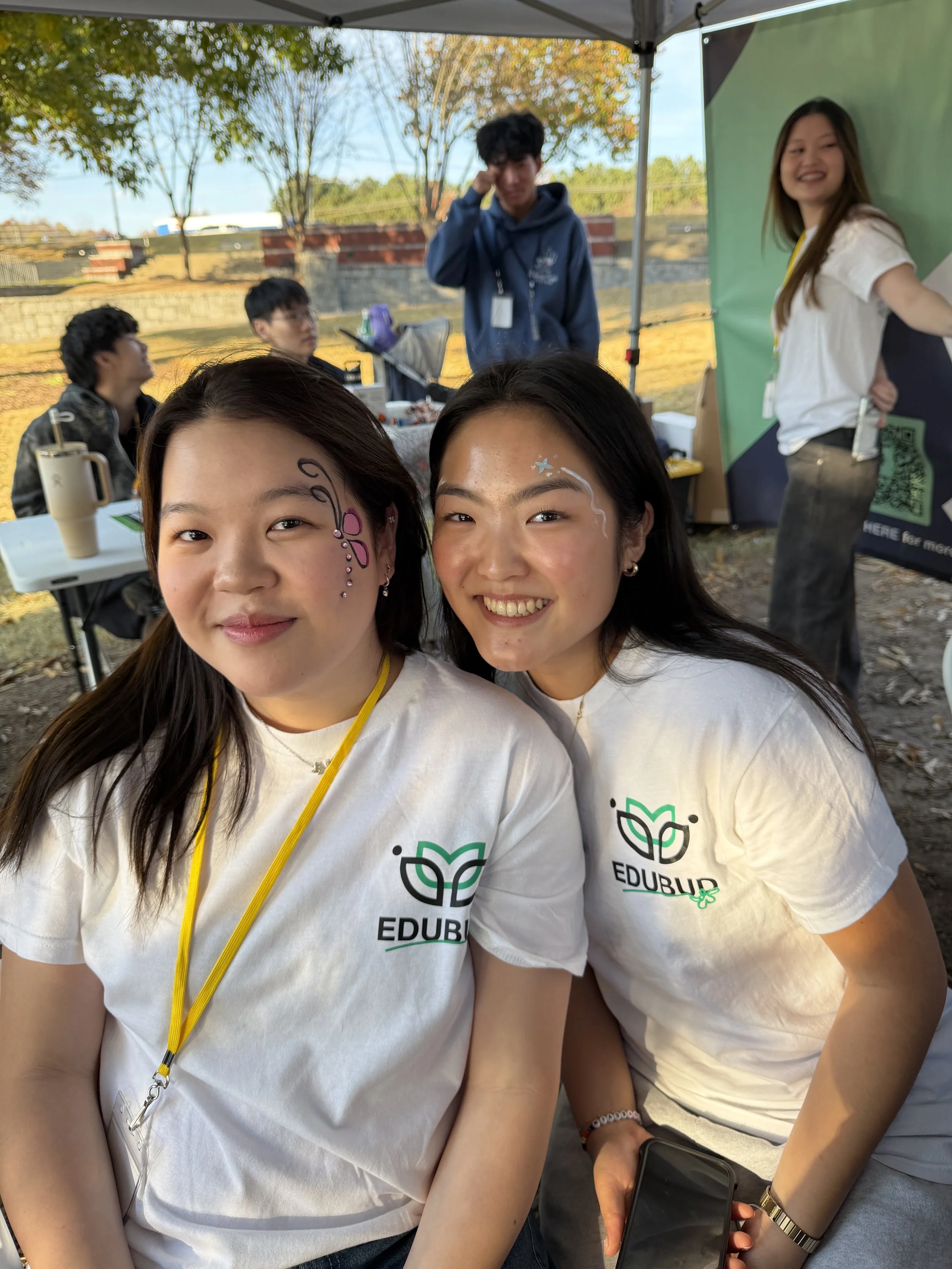 Two young women wearing white T-shirts with the Edbury logo, smiling and taking a selfie at an outdoor event. The background shows a tent, trees, and other people.