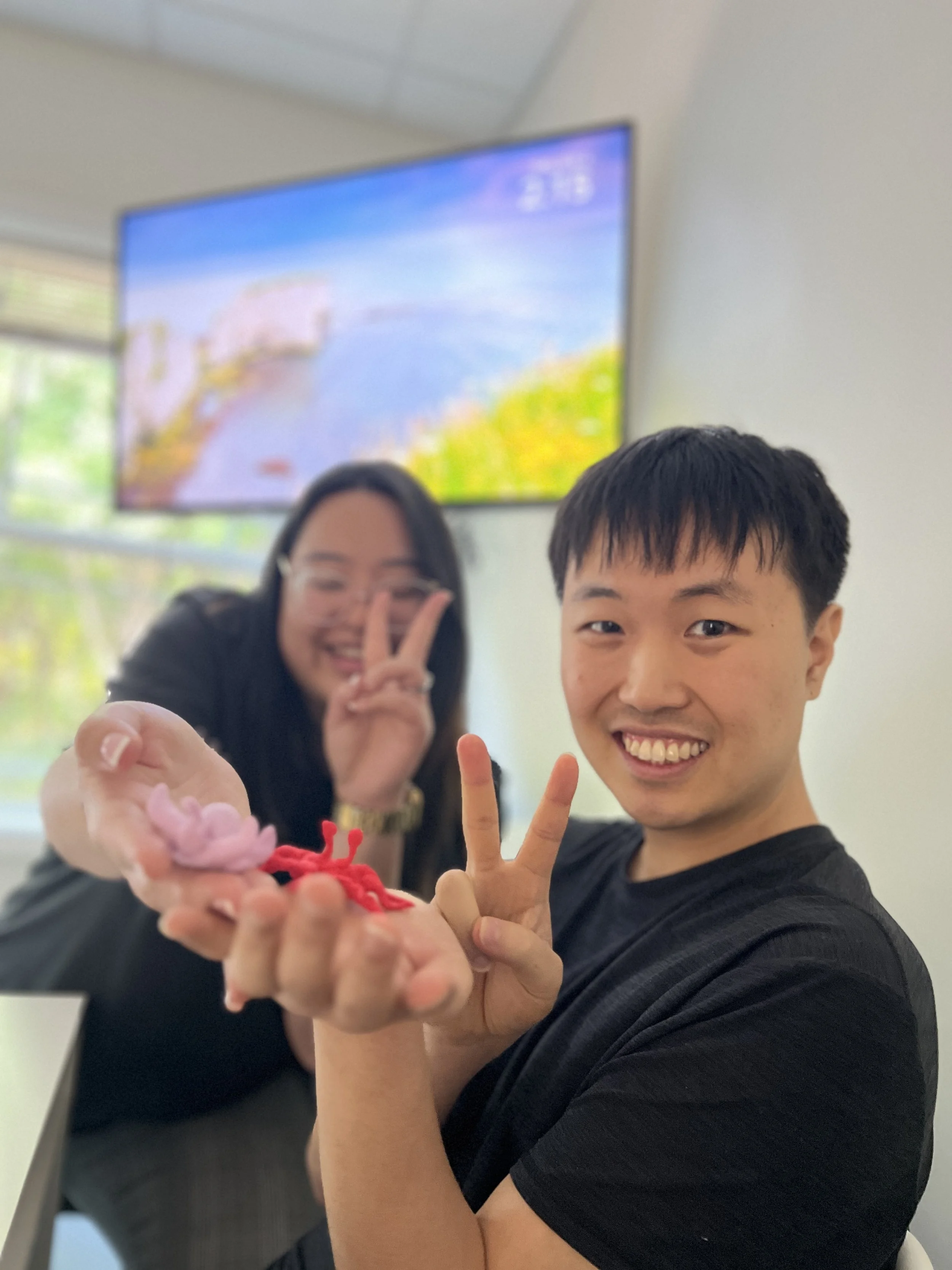Young man smiling and holding a small red and pink plush toy, with a woman in the background making a peace sign with her fingers, both in a room with a large window and a unsharp TV screen showing a landscape.