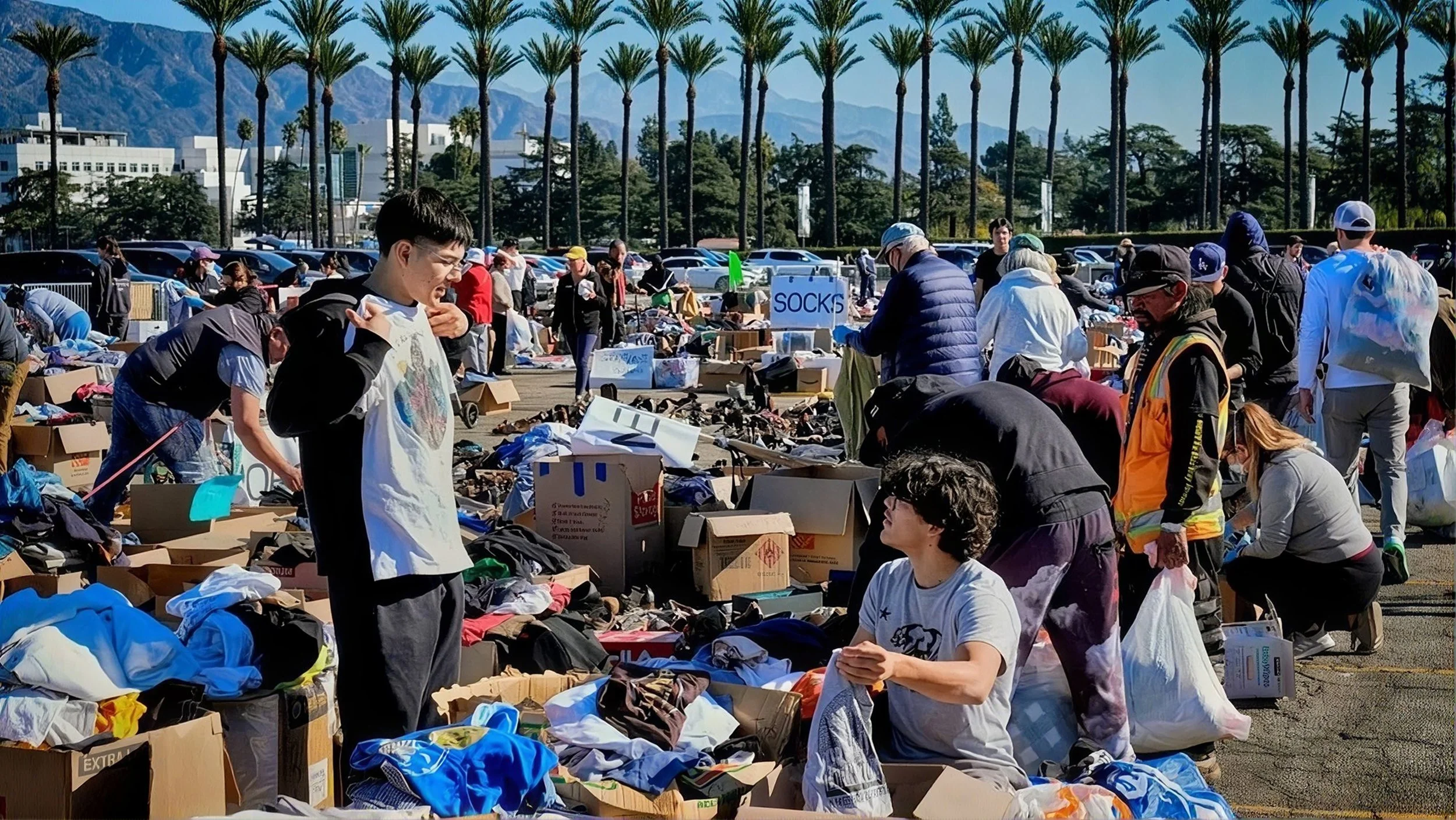 People browsing and shopping at an outdoor flea market with tables and boxes of secondhand items, trees and mountains in the background.