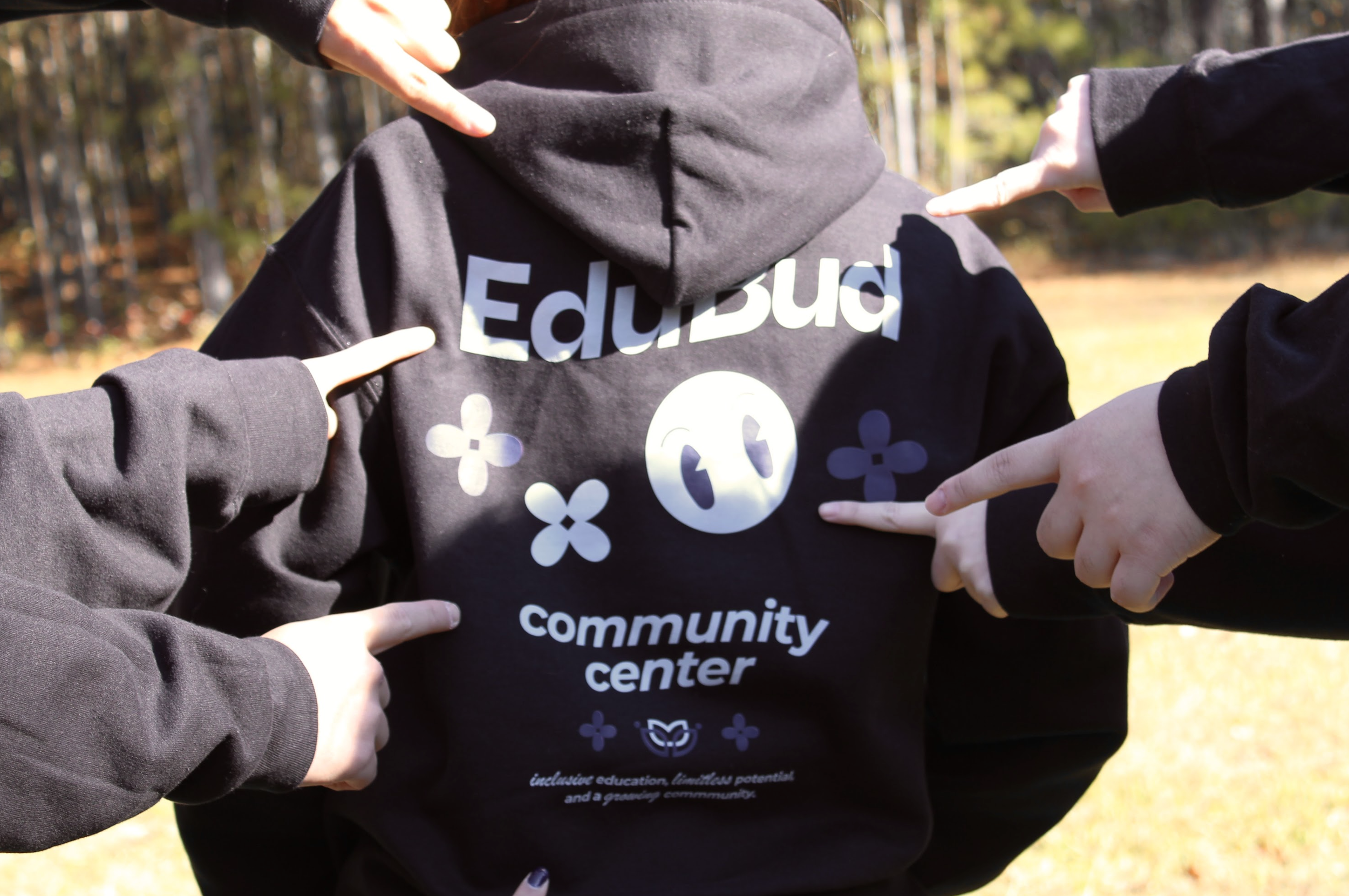 People pointing at a person wearing a black hoodie with 'EduBuc' printed on the back, outdoors in a park with trees in the background.