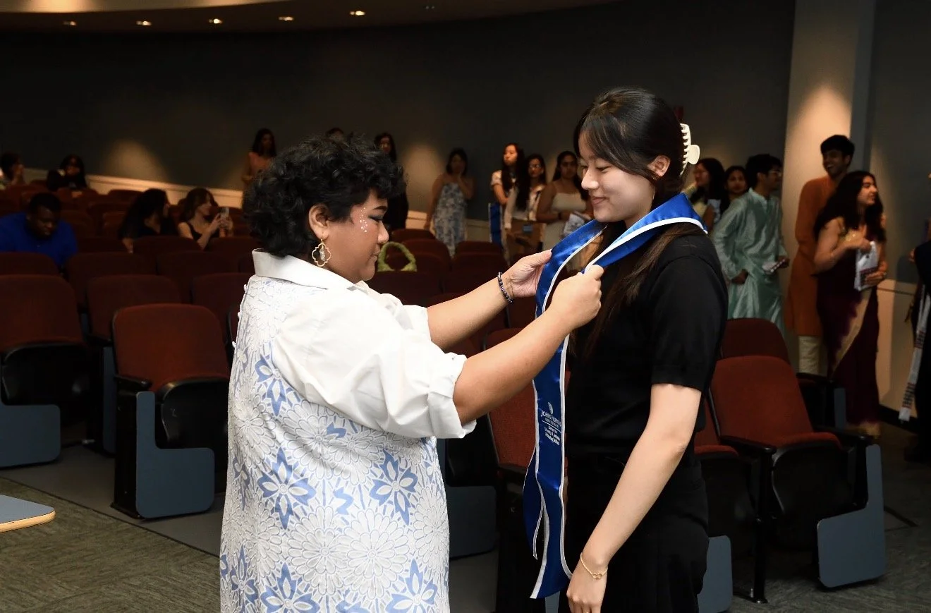 A woman in a white and blue floral dress awards a diploma or sash to a young woman dressed in black, in an auditorium with audience members seated and standing in the background.