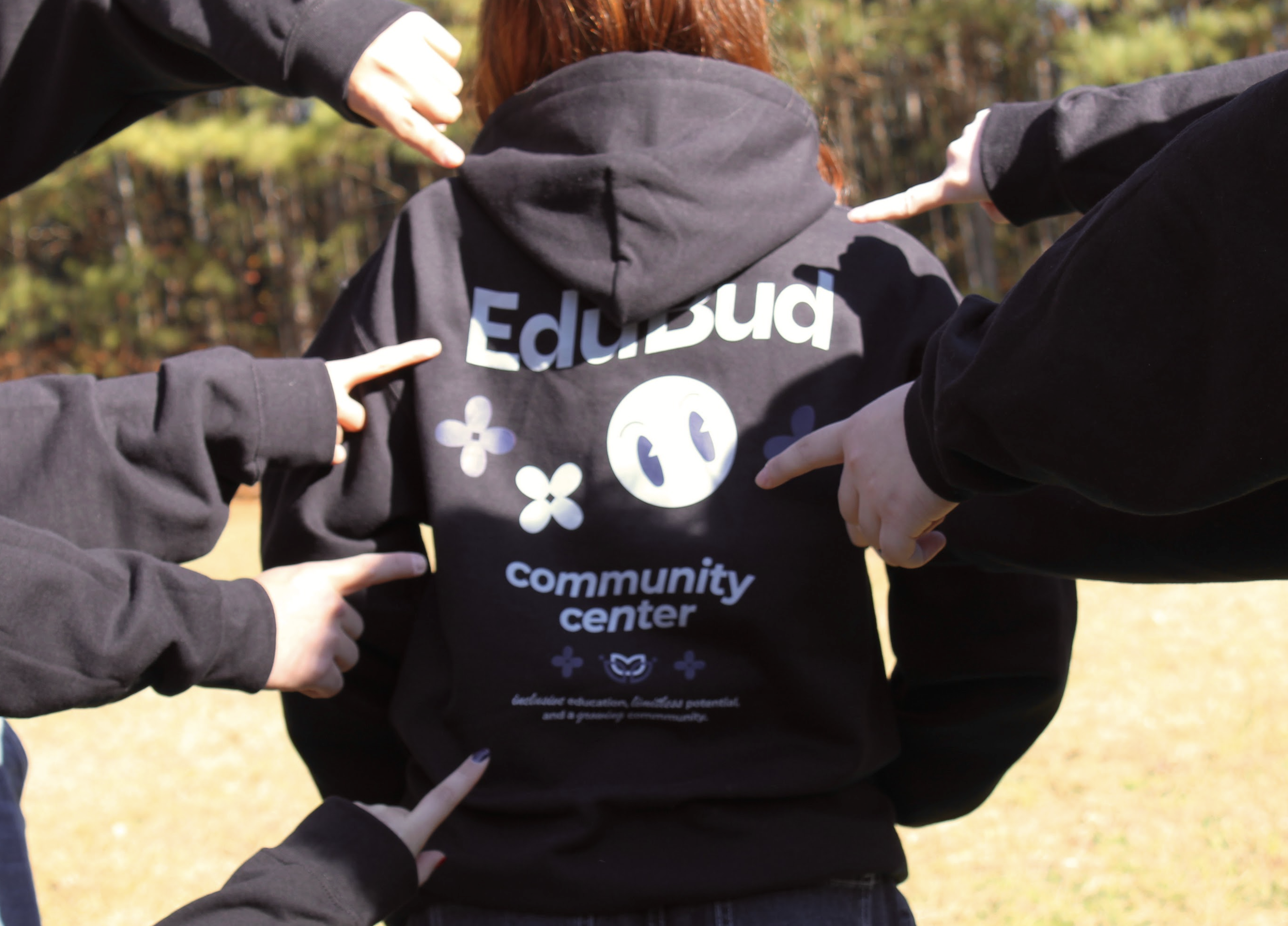 Group of people pointing towards a person wearing a black hoodie with the words 'Education', 'community center' and other text and symbols printed on the back, outside in a park or wooded area with trees in the background.