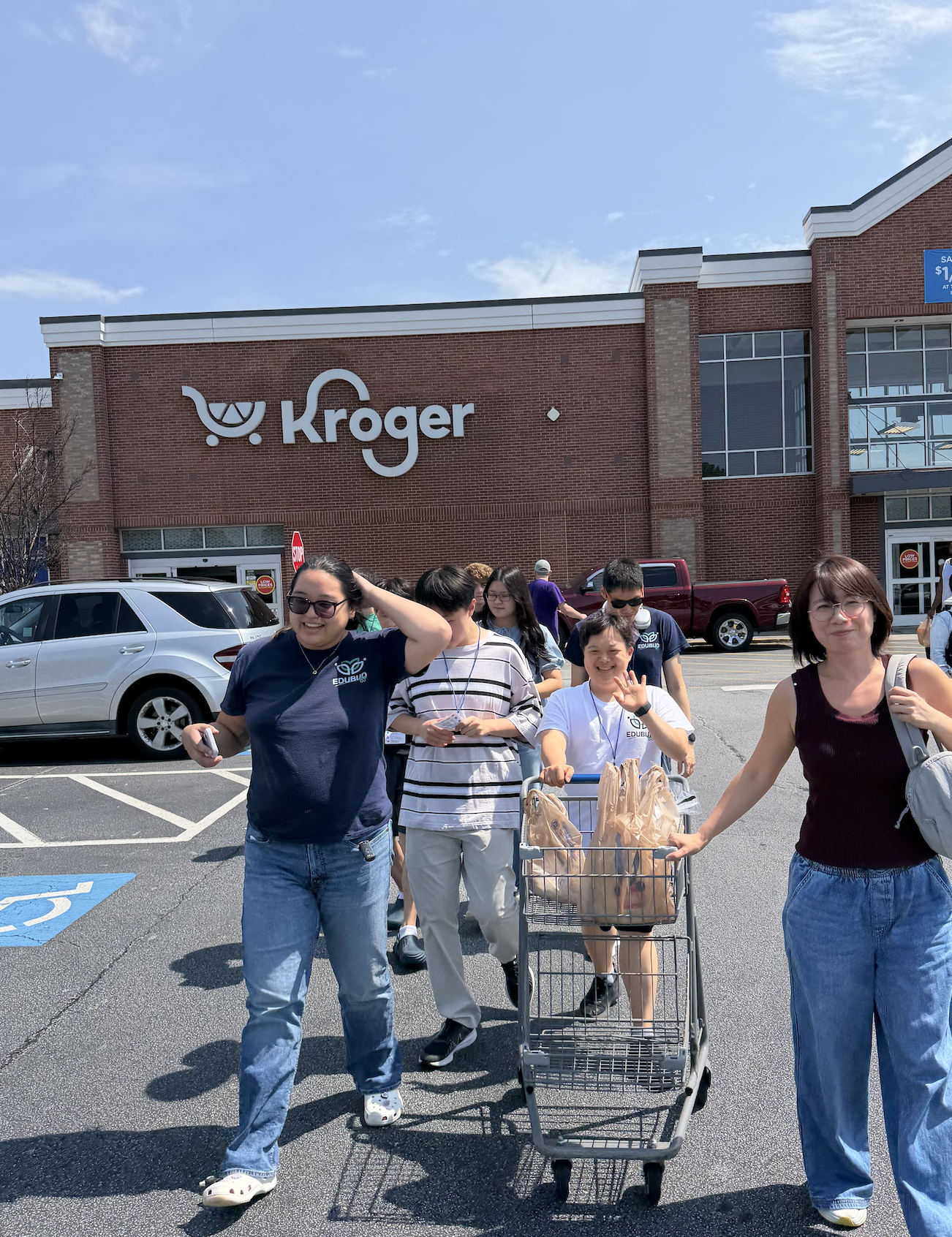 People walking out of a Kroger grocery store, some smiling and pushing a shopping cart filled with grocery bags.