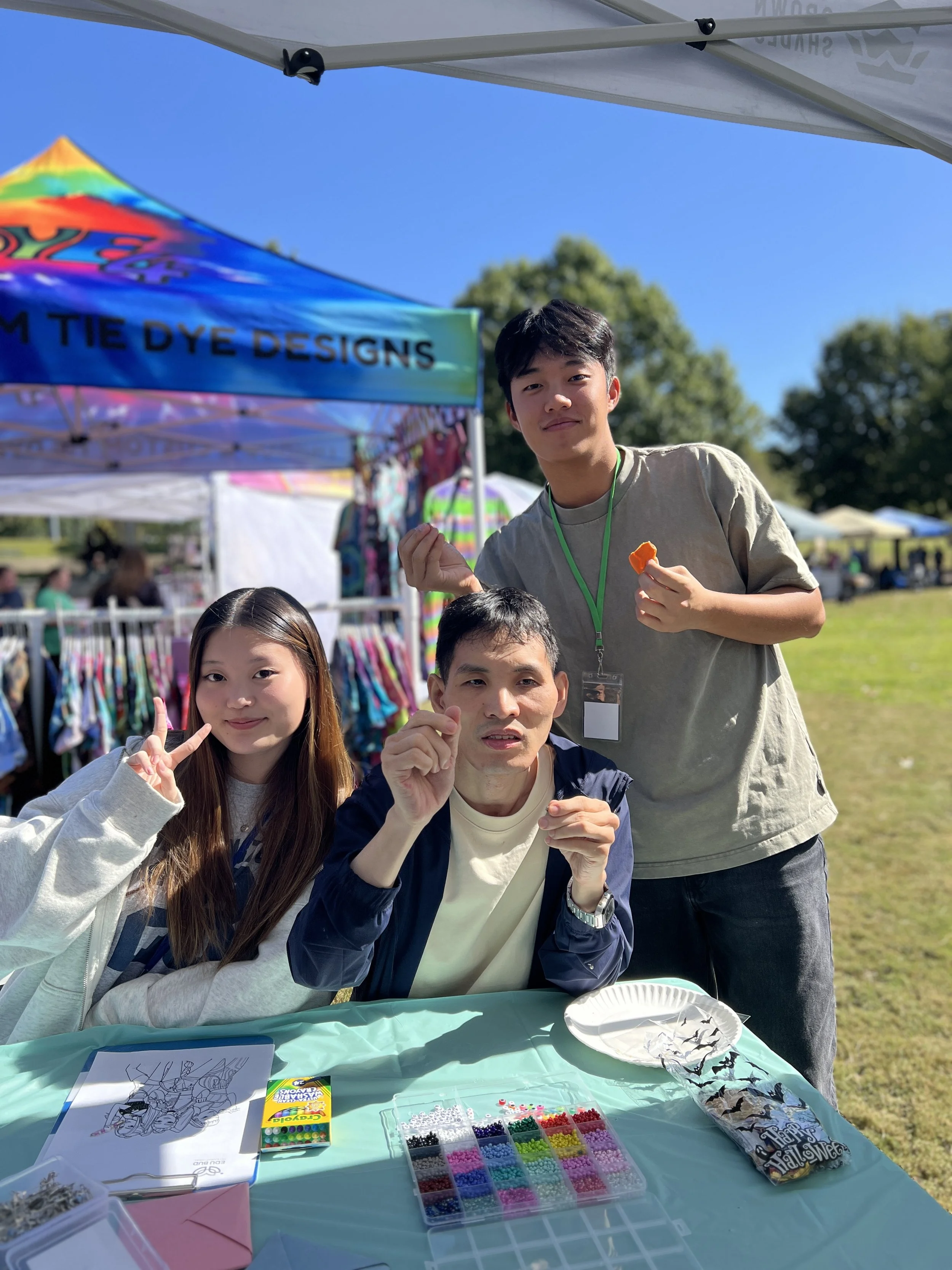 A group of people at an outdoor event under a white canopy, engaging in arts and crafts. There are craft supplies on a table including beads and coloring sheets, with a colorful rainbow-themed banner in the background.