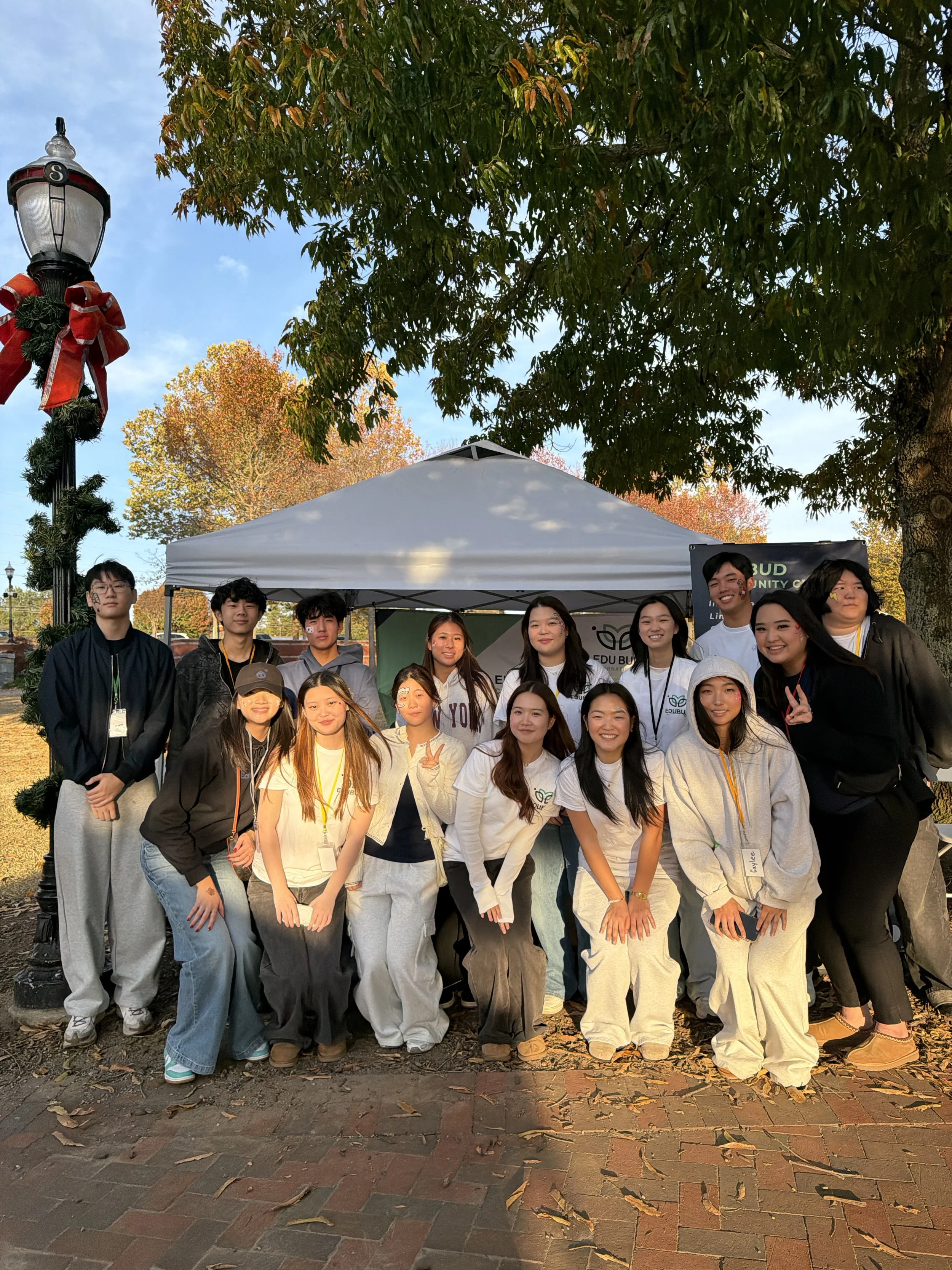 A group of young people, mostly women, standing outdoors under trees with fall foliage, in front of a white tent during the daytime.