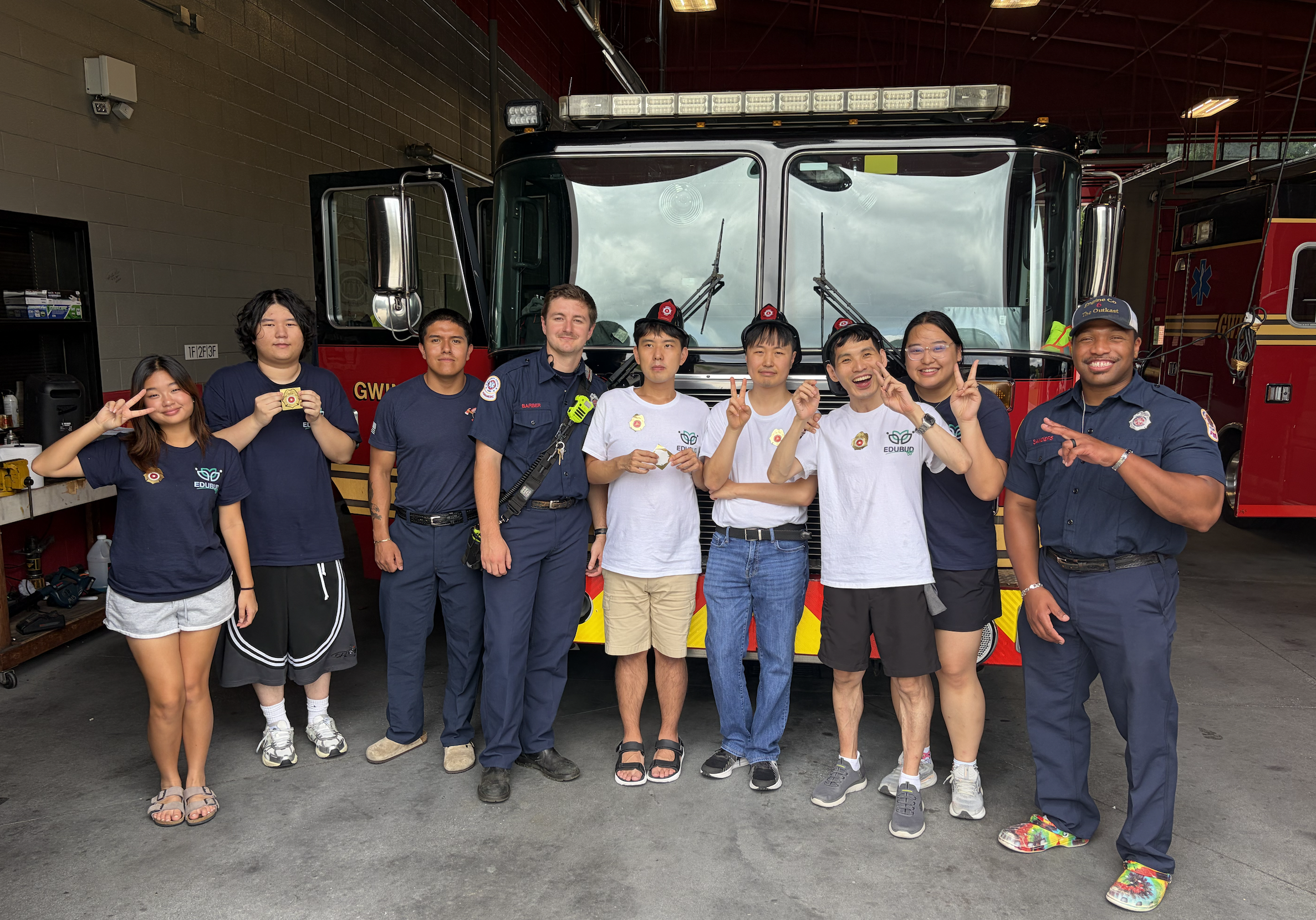 A group of nine people, including individuals in firefighter uniforms and casual clothing, standing in front of a fire truck inside a garage or fire station. Some are making peace signs and smiling.