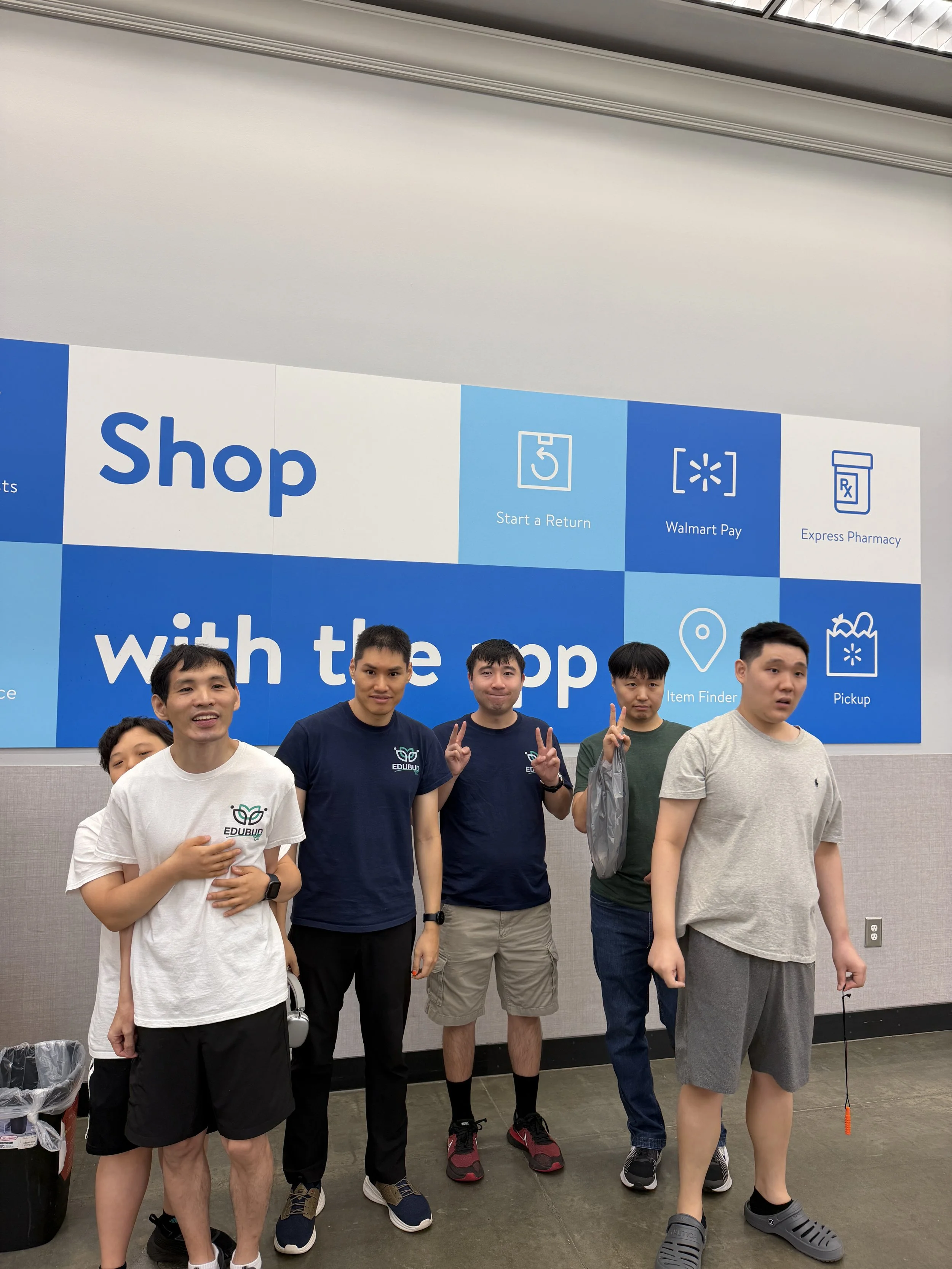Group of five young men standing indoors in front of a large blue sign with instructions for shopping, returning, Walmart Pay, pharmacy, and item pickup.