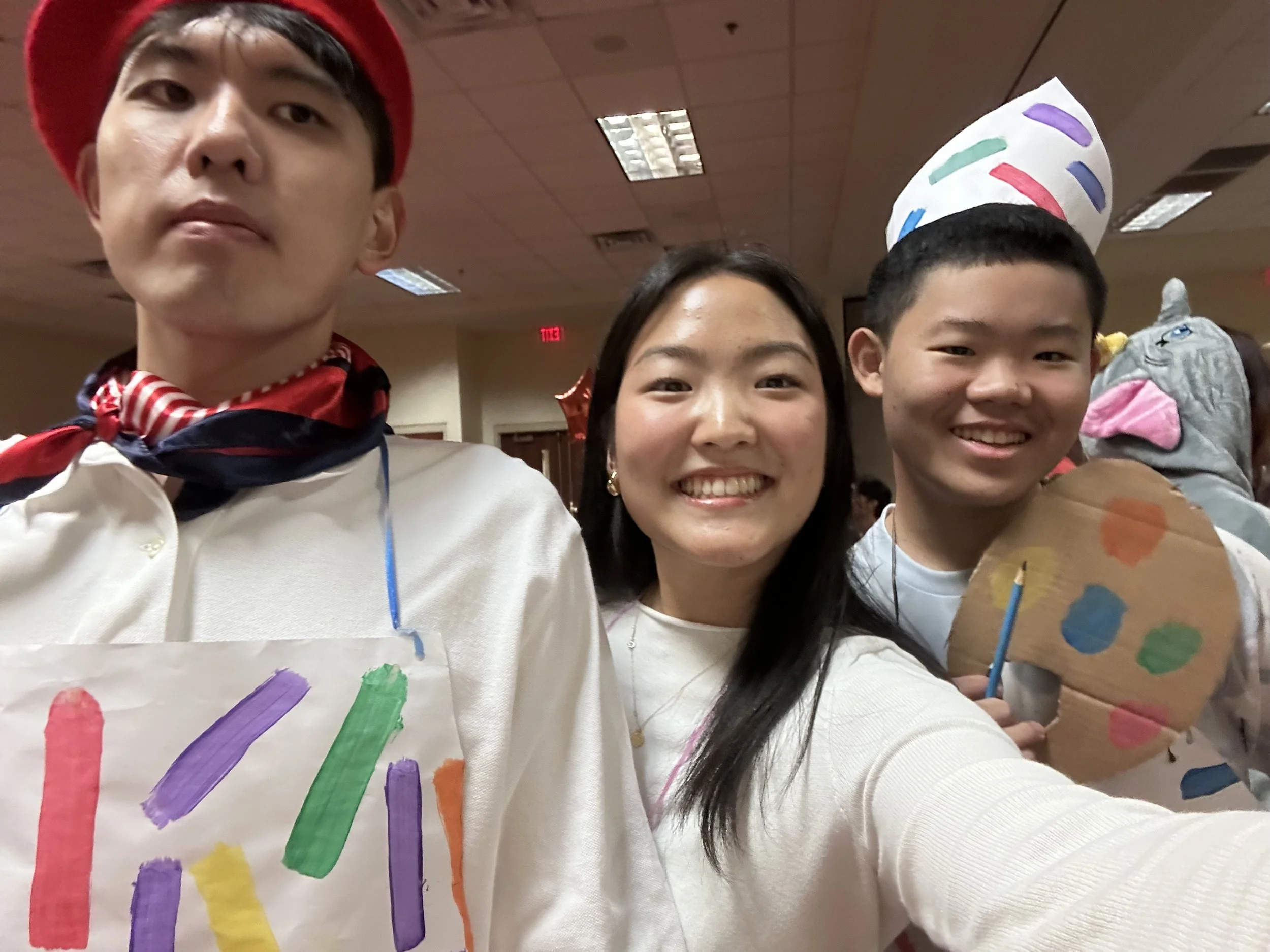 Three young individuals celebrating a birthday or special occasion in a party room, wearing paper hats and colorful shirts with painted rainbow designs, smiling and taking a group selfie.