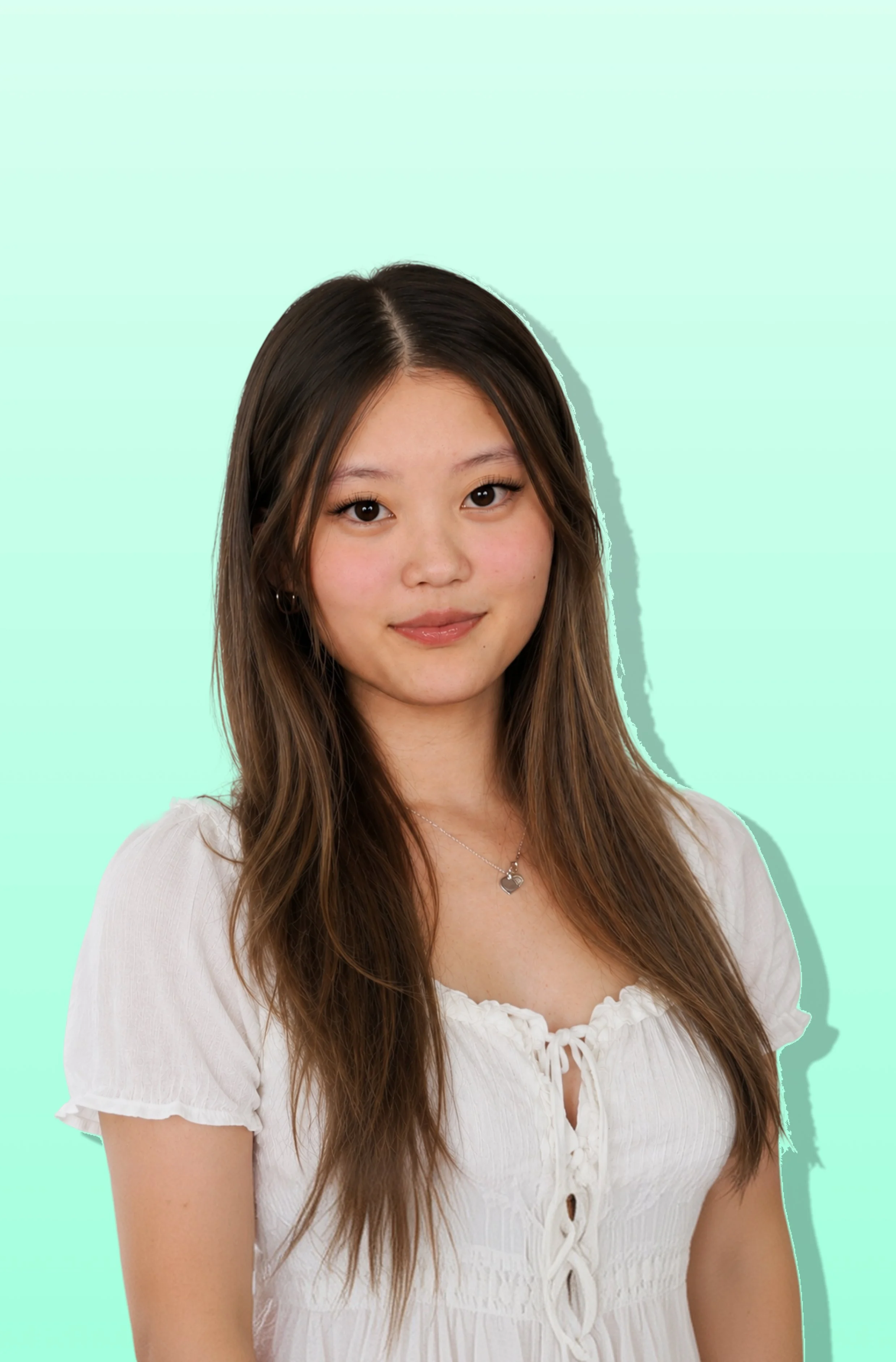 Close-up of a young woman with long brown hair, wearing a white blouse and a heart-shaped necklace, standing against a mint green background.