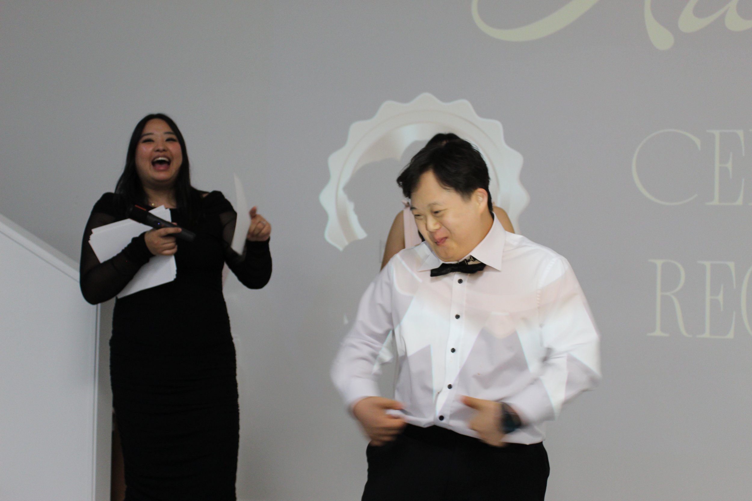 Two people, a woman and a man, are at an event. The woman is wearing a black dress and holding papers and a microphone, laughing. The man is in a white shirt with a black bowtie, looking down and smiling. They are standing in front of a plain wall wi