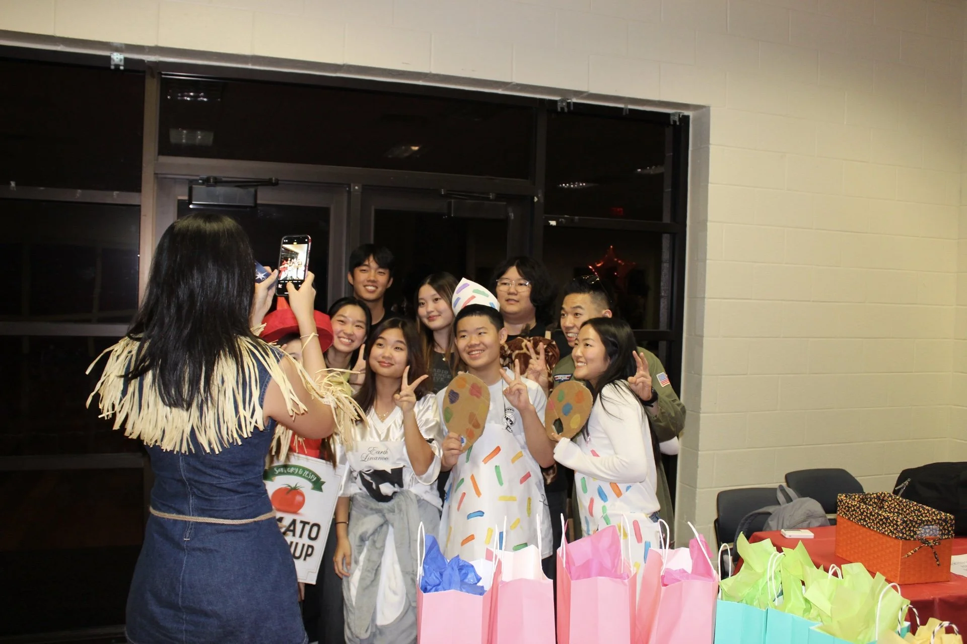 Group of children and teenagers dressed in costumes, taking a group photo at a celebration with gift bags on the table in front.