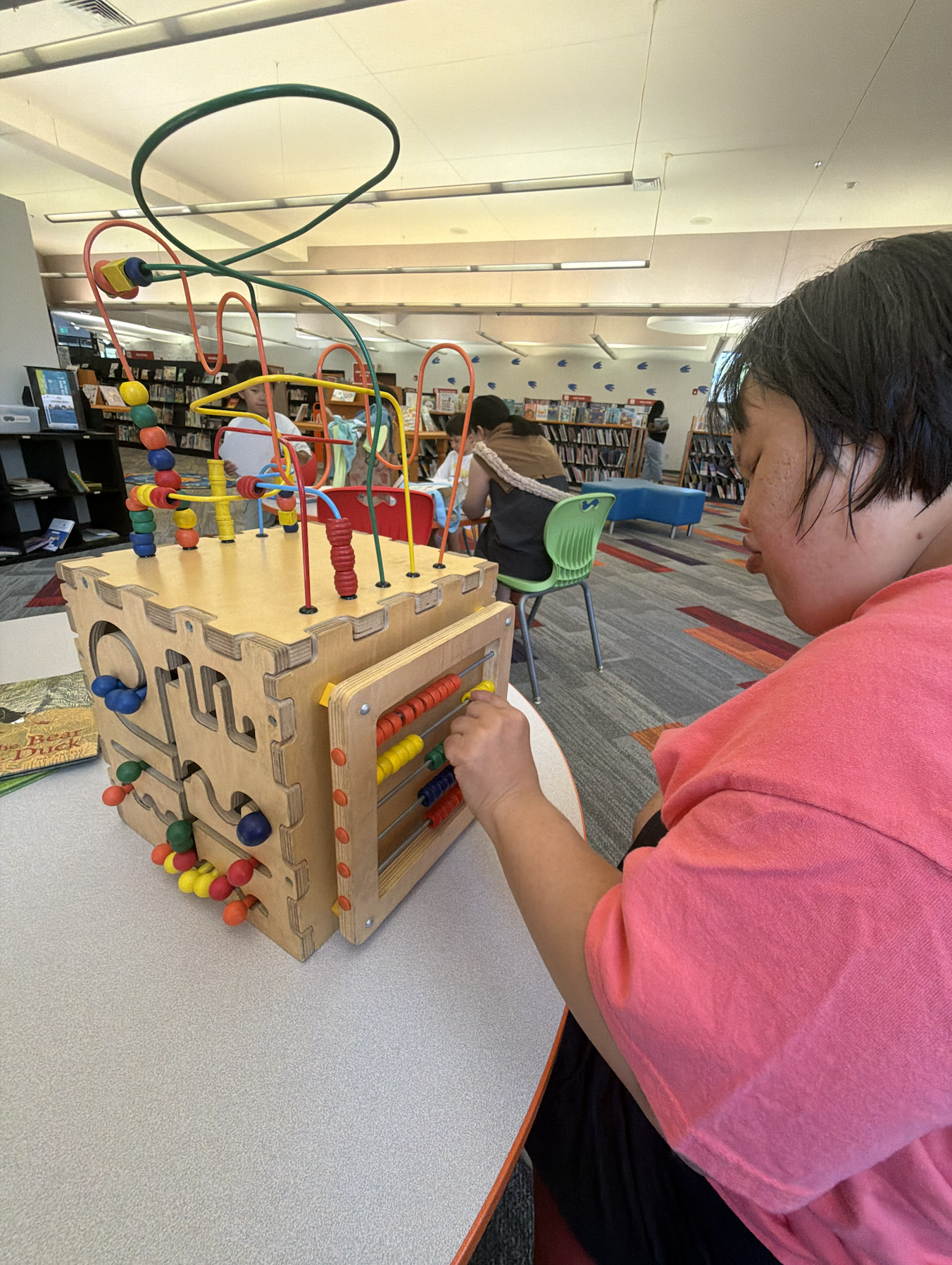 A young girl with short dark hair and a pink shirt plays with a wooden bead maze toy at a library. In the background, there are people sitting at tables and browsing bookshelves.