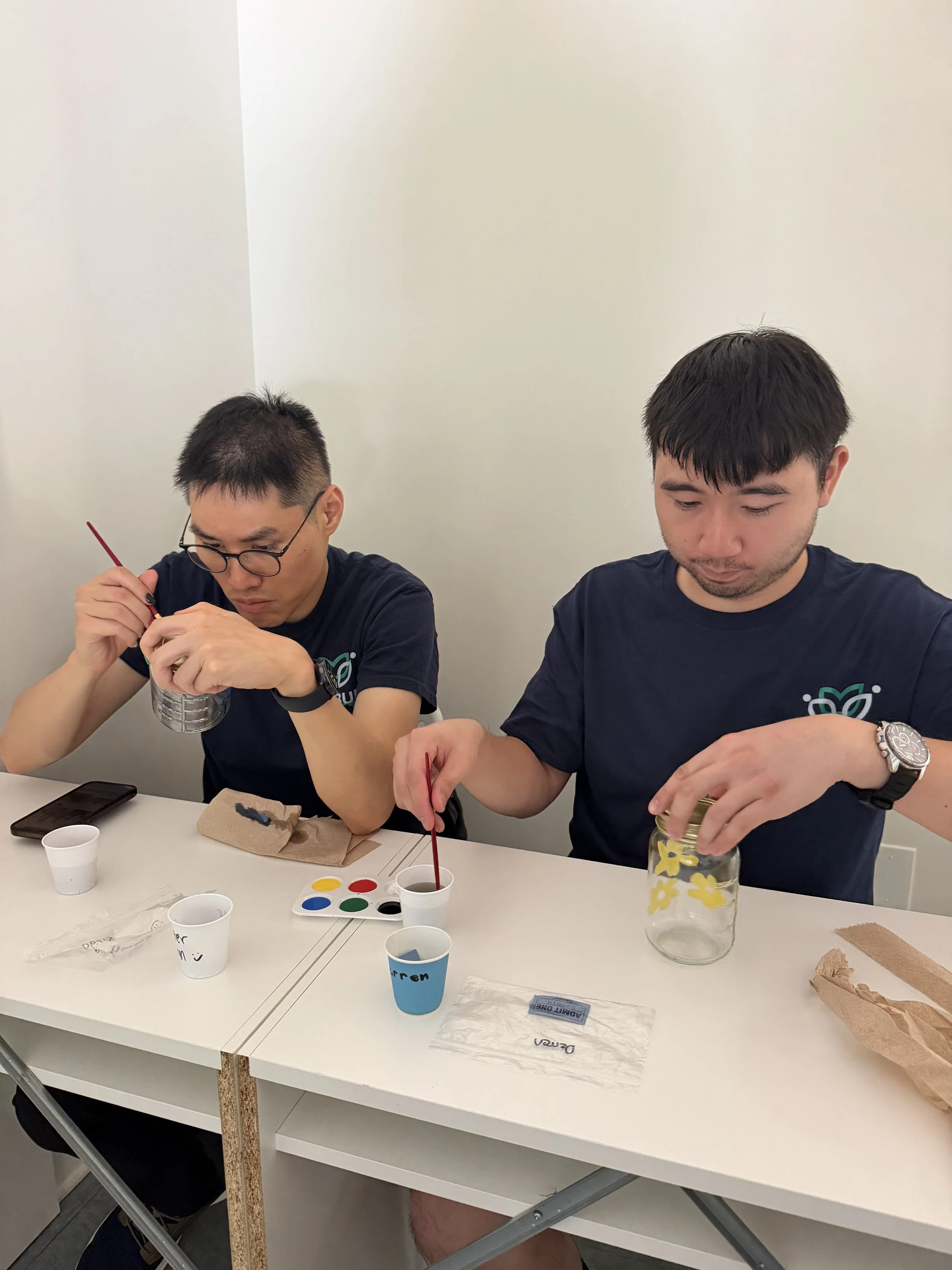 Two men sitting at a white table, working on a craft project with small cups of colored paint, a glass jar, and paper towels. One man is wearing glasses and a black shirt, the other has dark hair and a watch.
