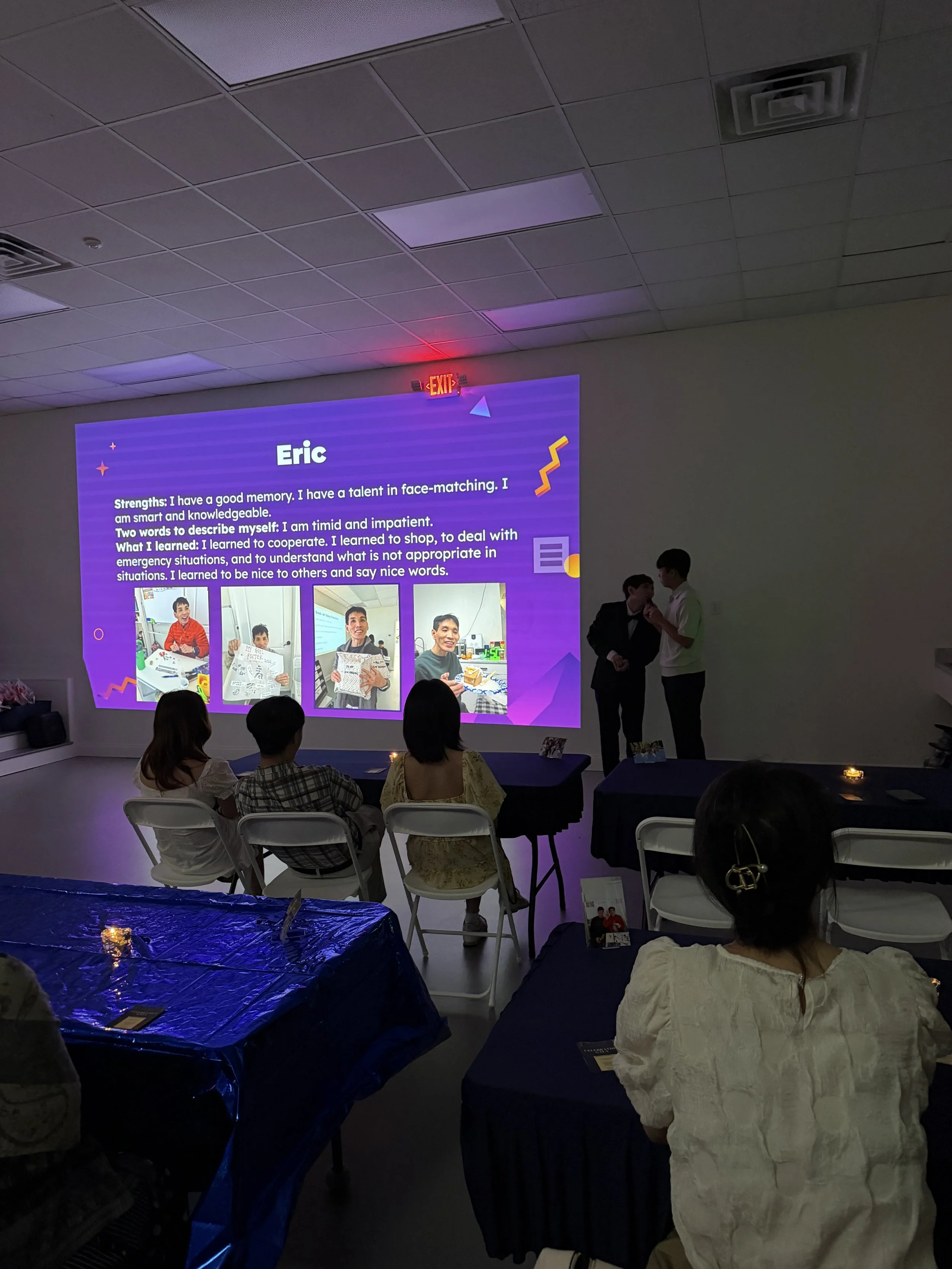 A group of people attending an indoor presentation with a large screen displaying a biography of Eric, including his strengths and personal development points. The room has a white ceiling with fluorescent lights and some people are standing while ot