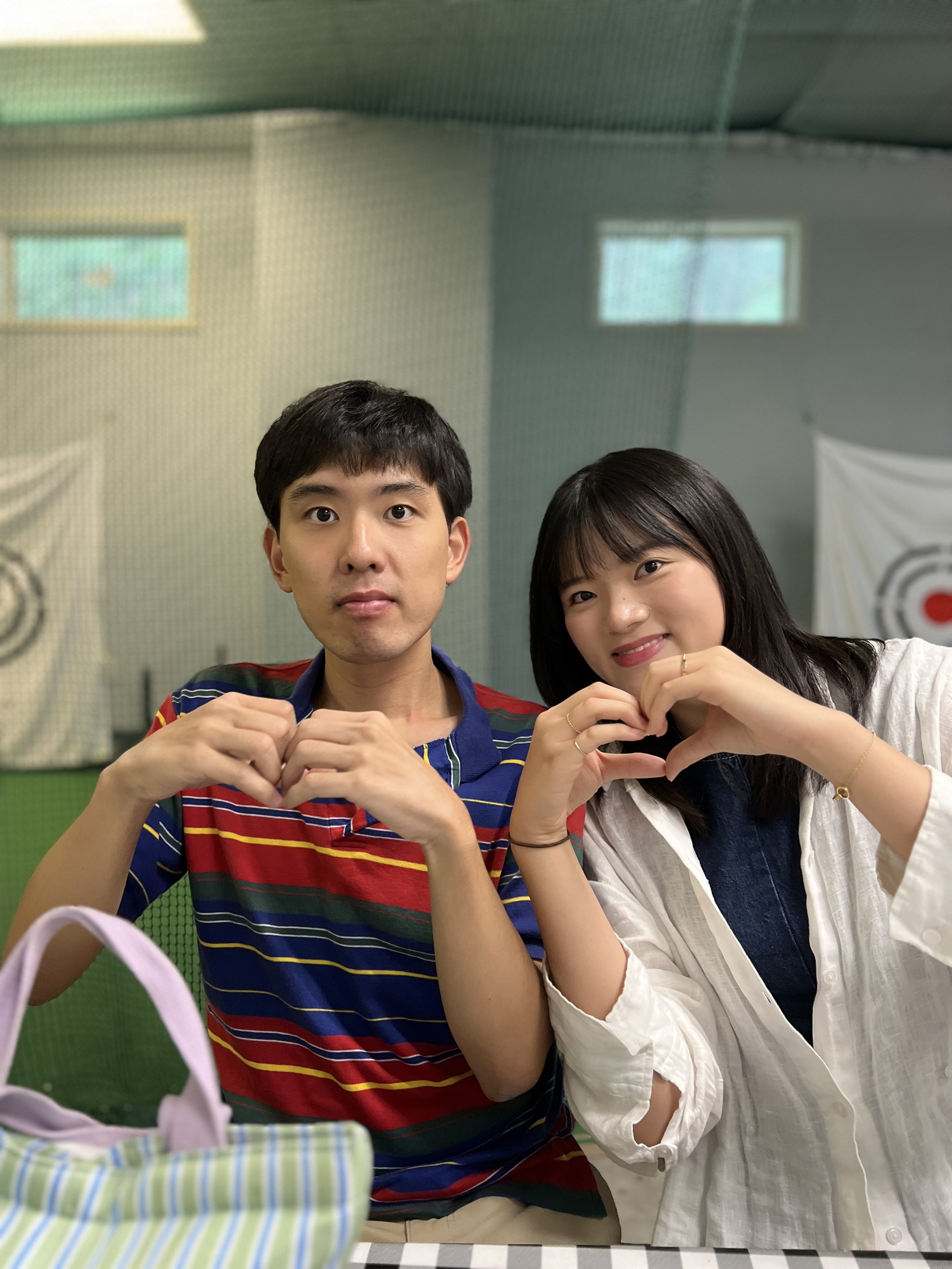 A young man and woman sitting together at a table, making heart shapes with their hands, smiling, in a room with green walls, windows, and a partially visible flag.
