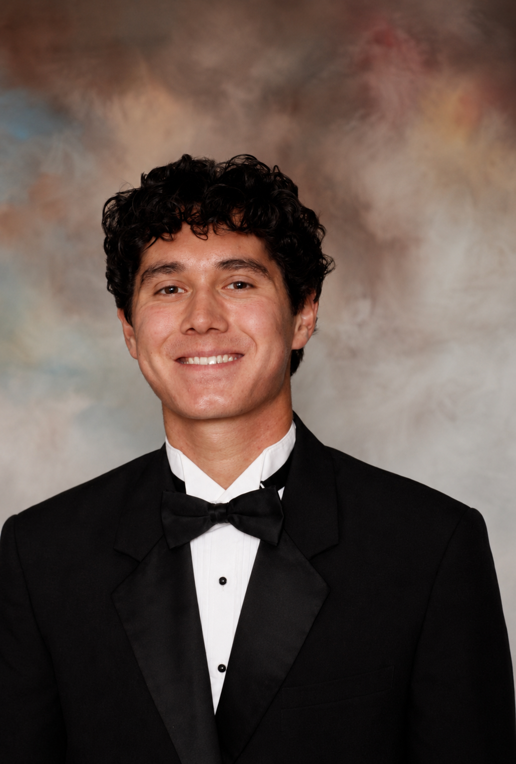 A young man dressed in a black tuxedo and bowtie, smiling at the camera against a multicolored blurred background.