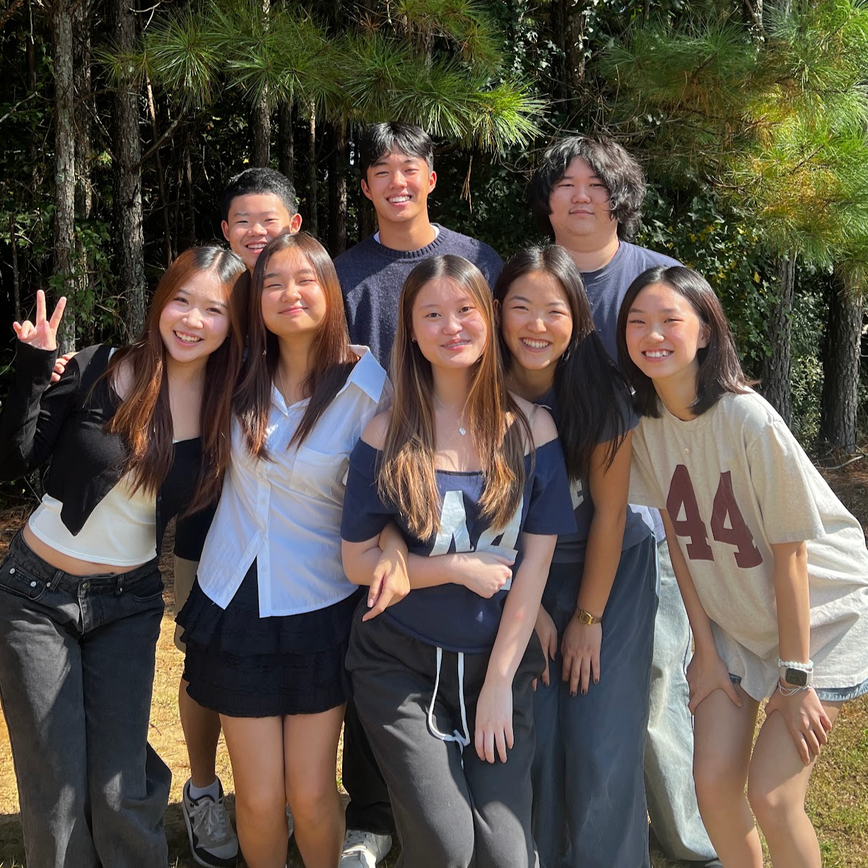 A group of nine young friends smiling and posing outdoors in front of trees. They are dressed casually, with one making a peace sign.