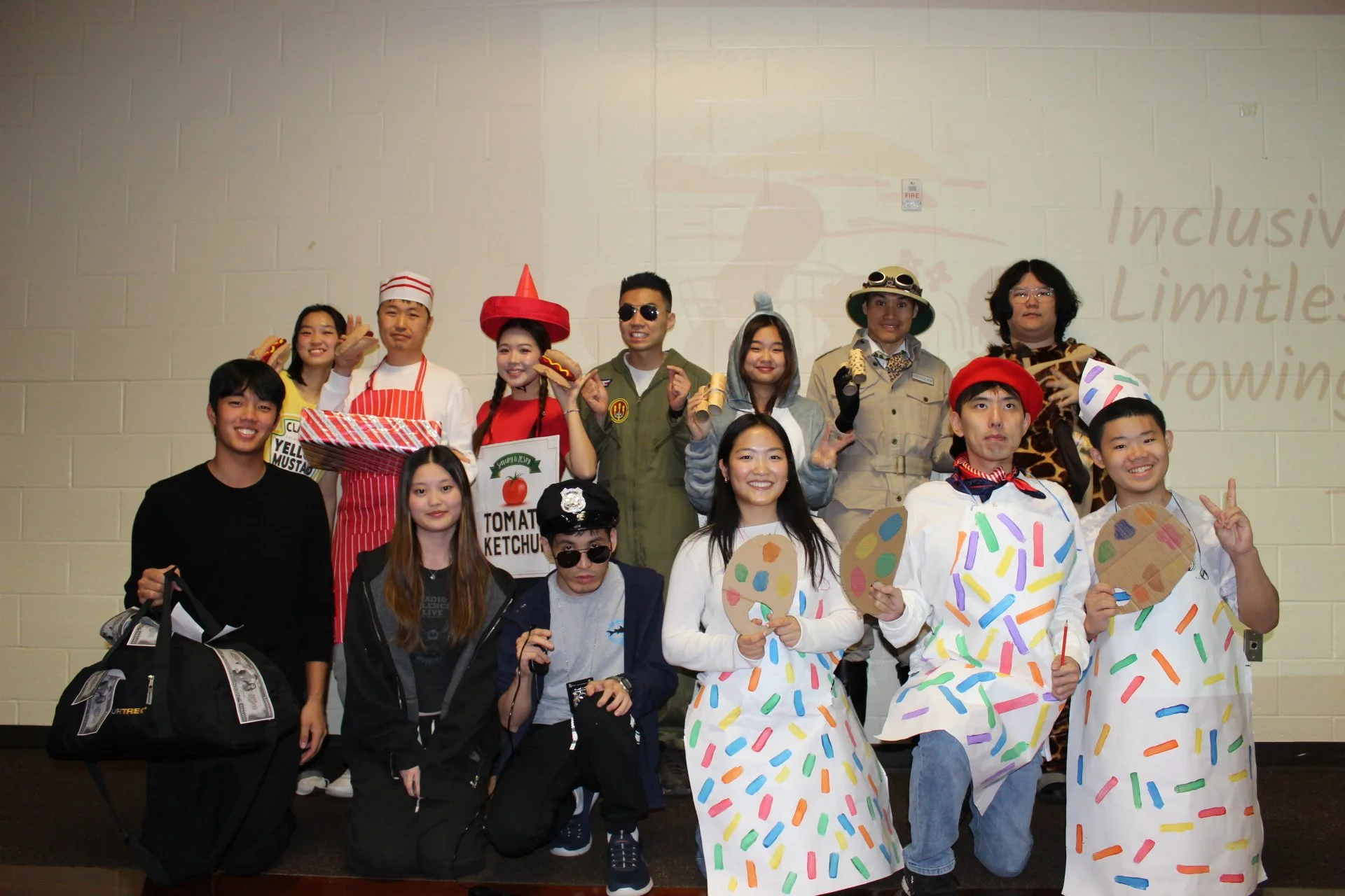 Group of diverse people dressed in costumes and casual clothing, some holding food and props, standing in front of a beige brick wall with partially visible words 'Inclusive' and 'Limitless' and a fire alarm.