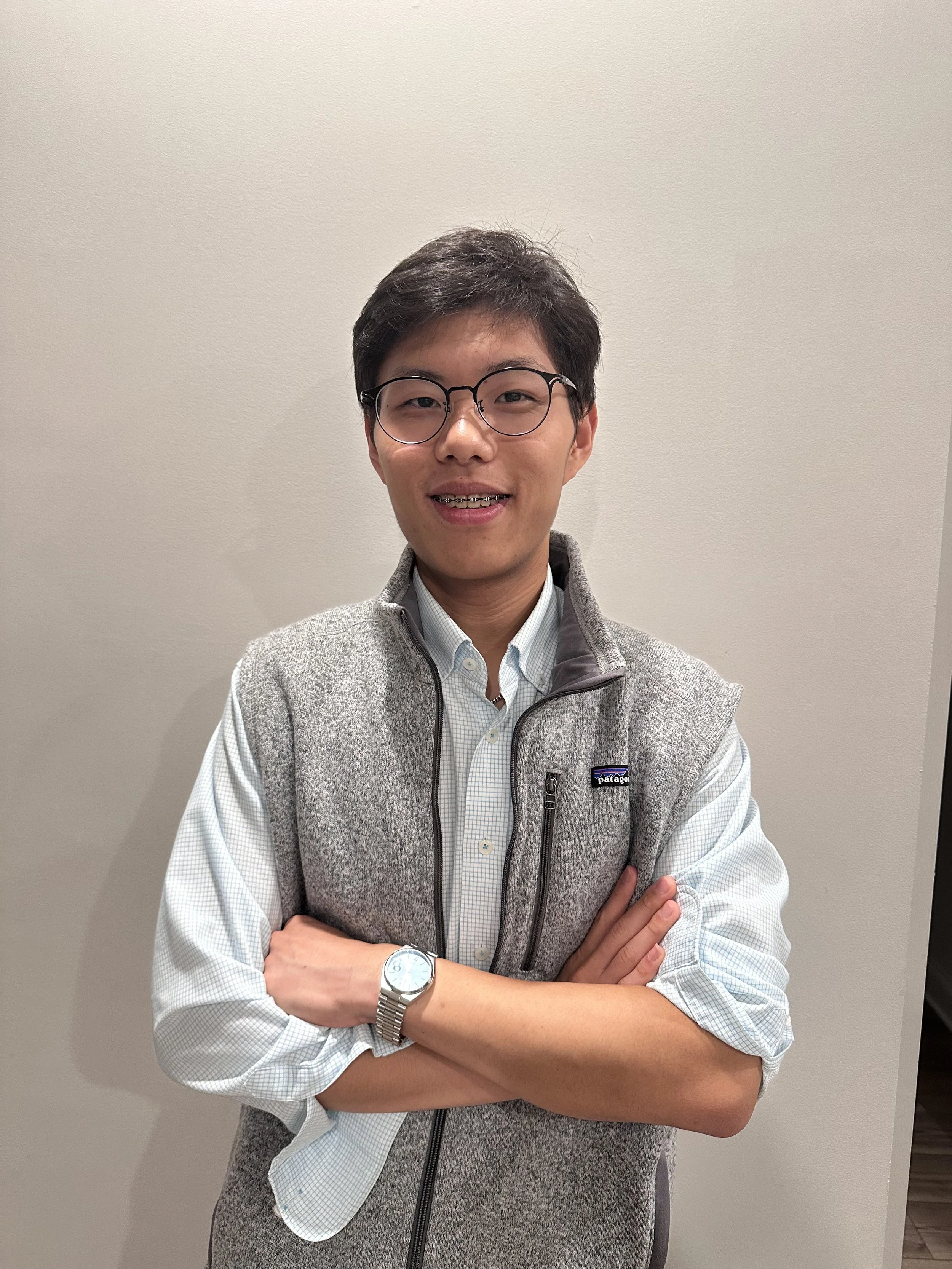 A young man with short dark hair, glasses, and a light-colored shirt, smiling with his arms crossed, standing against a plain beige wall.