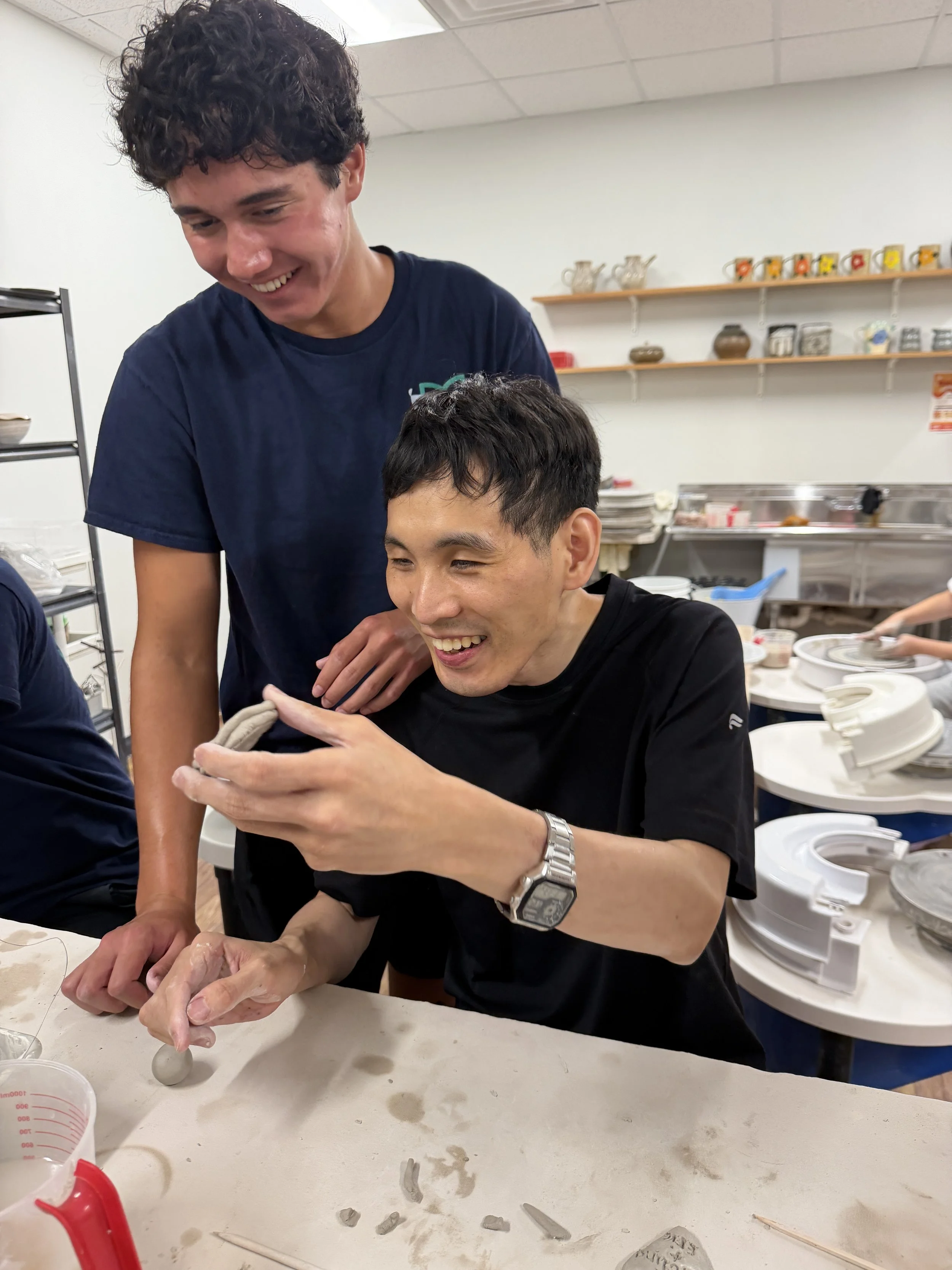 Two young men are making pottery together in a ceramics class, smiling and enjoying the activity in a bright room with shelves holding various ceramic bowls and decorative items.