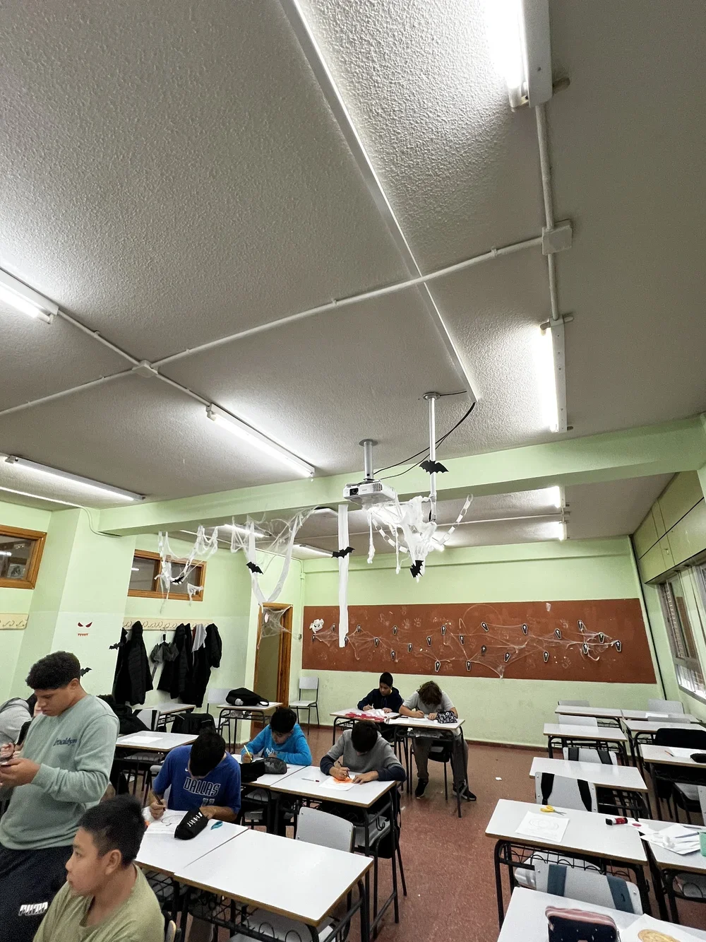 Classroom decorated with Halloween decorations, including white cobwebs and black bats hanging from the ceiling. Students are seated at desks, some working and some standing, in a room with green walls and large windows.