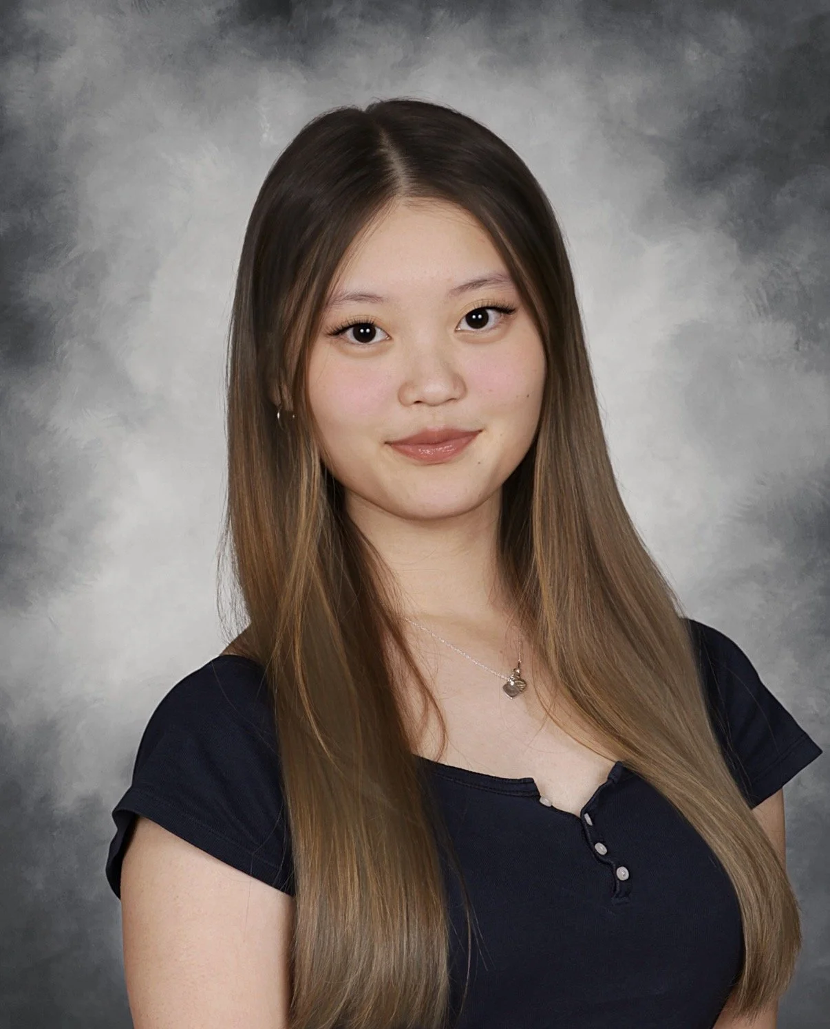 Portrait of a young woman with long brown hair, wearing a navy blue top and a silver necklace, standing against a gray mottled background.