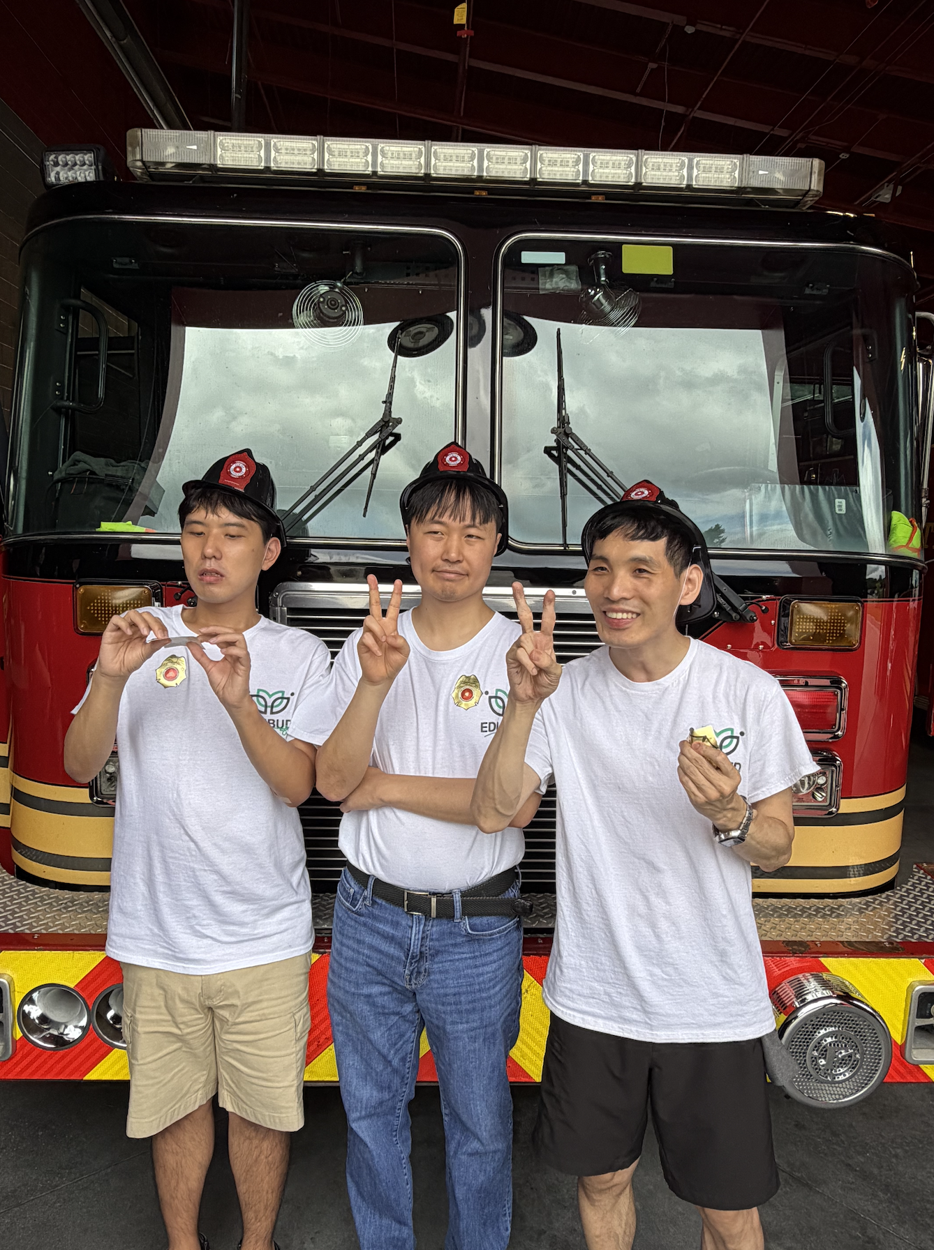 Three men standing in front of a fire truck posing for a photo. Two of the men are making peace signs with their hands, and all three are holding small items in their hands. They are wearing white T-shirts and fire helmets.